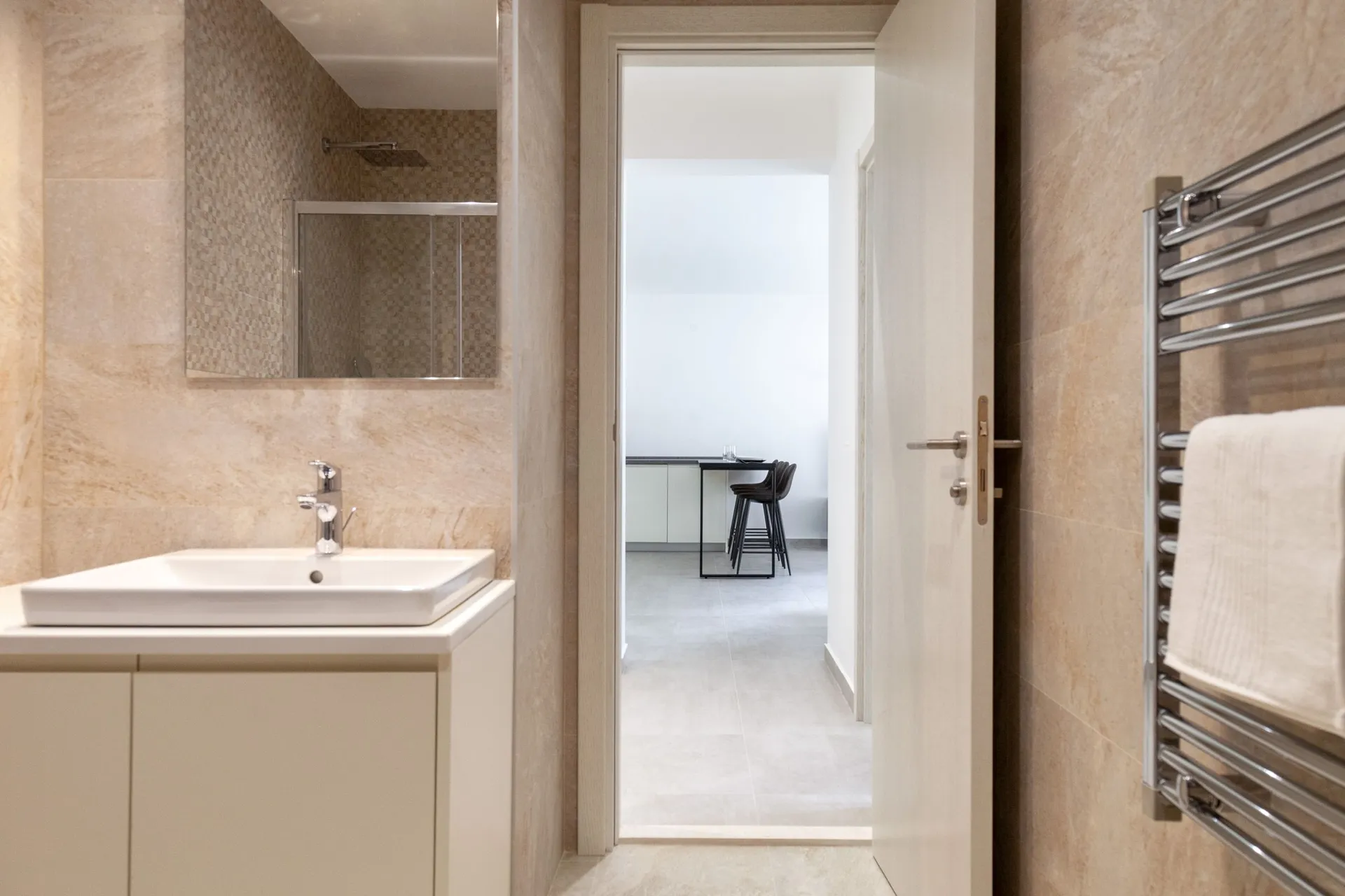 Modern bathroom with beige tiles, a white vanity sink, and a chrome heated towel rail visible on the right wall.
