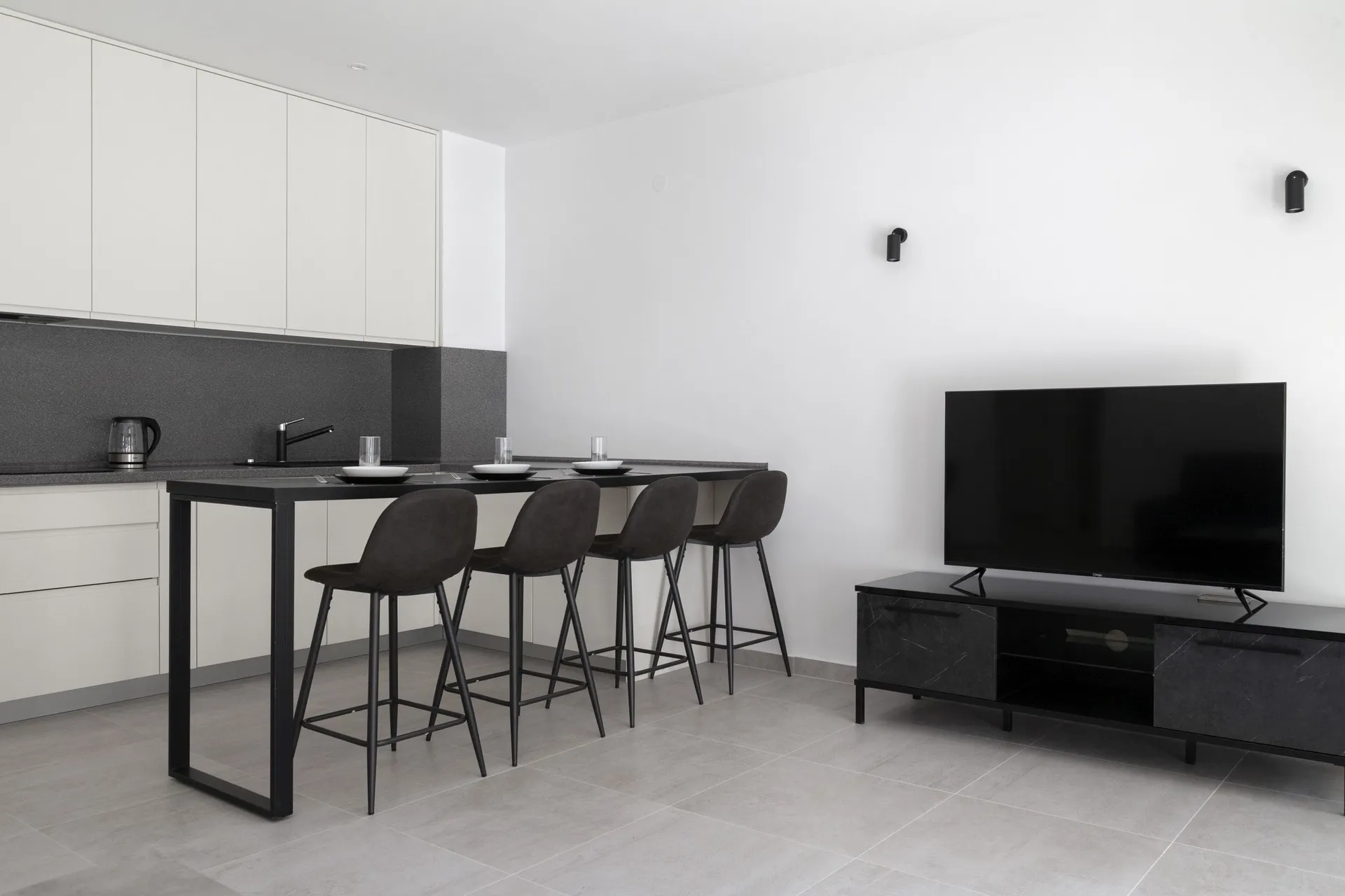 Modern kitchen with white cabinets, black countertop, bar stools, and a TV on a dark media console.
