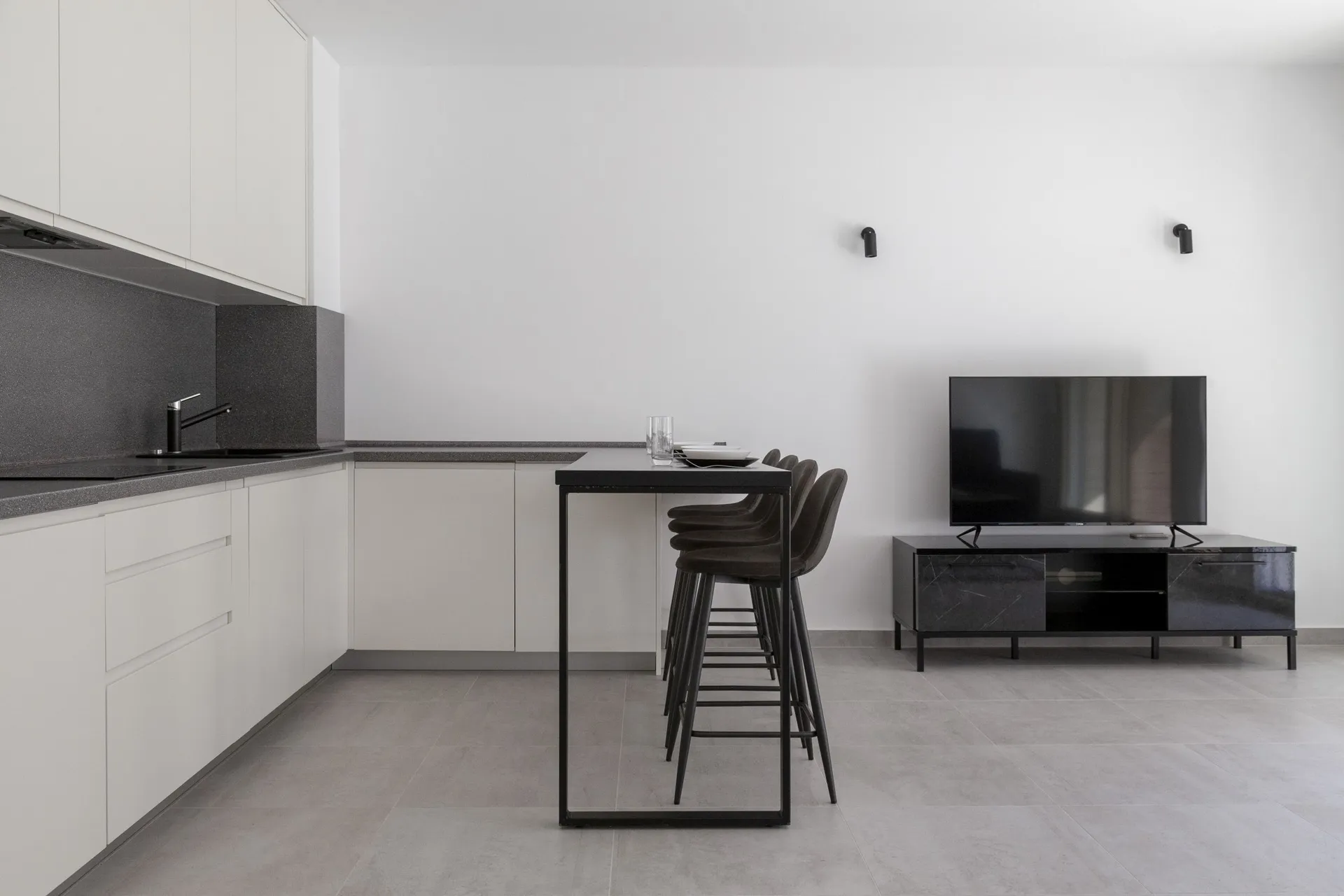 Modern kitchen with white cabinets, black countertop, bar table with stools, and a television on a dark media console.
