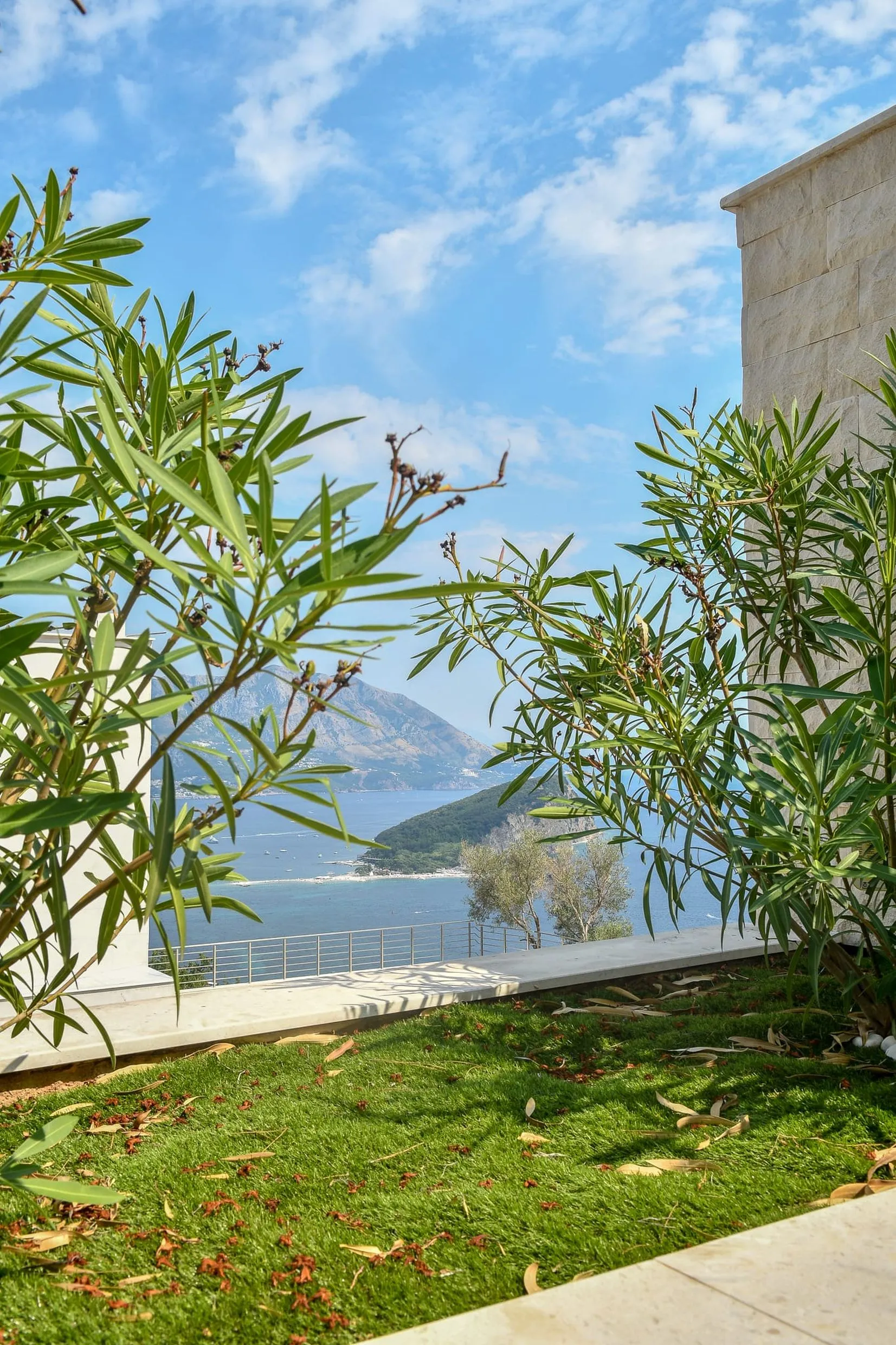 Sea and mountain view from a terrace with green grass, shrubs, stone wall, and blue sky.