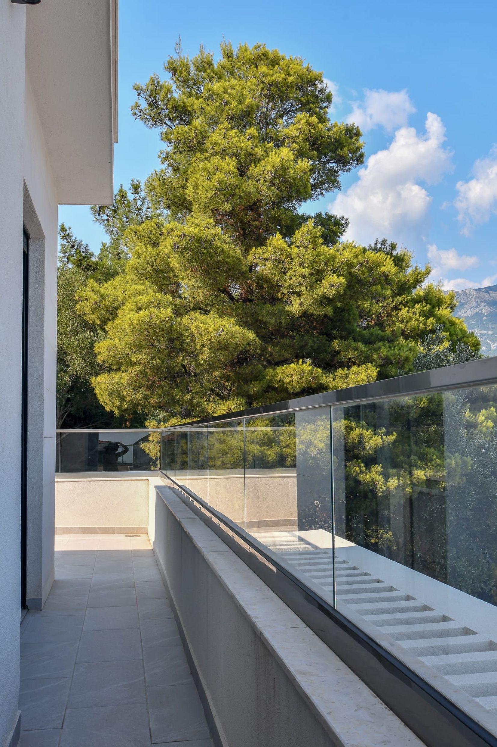 Spacious balcony with panoramic glass railing and a view of conifer trees at a villa in Budva, Montenegro.