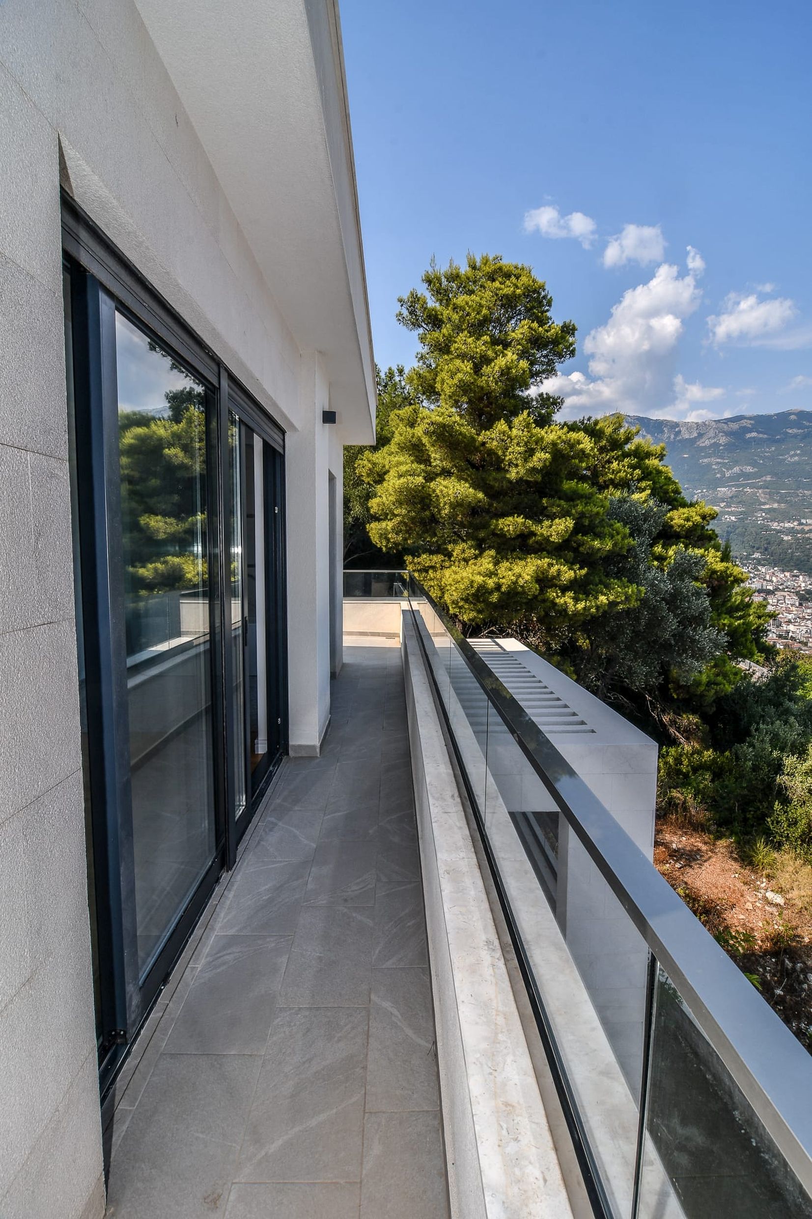 Modern villa balcony in Budva, Montenegro, with panoramic glazing, stone flooring, and views of the mountains and sea.