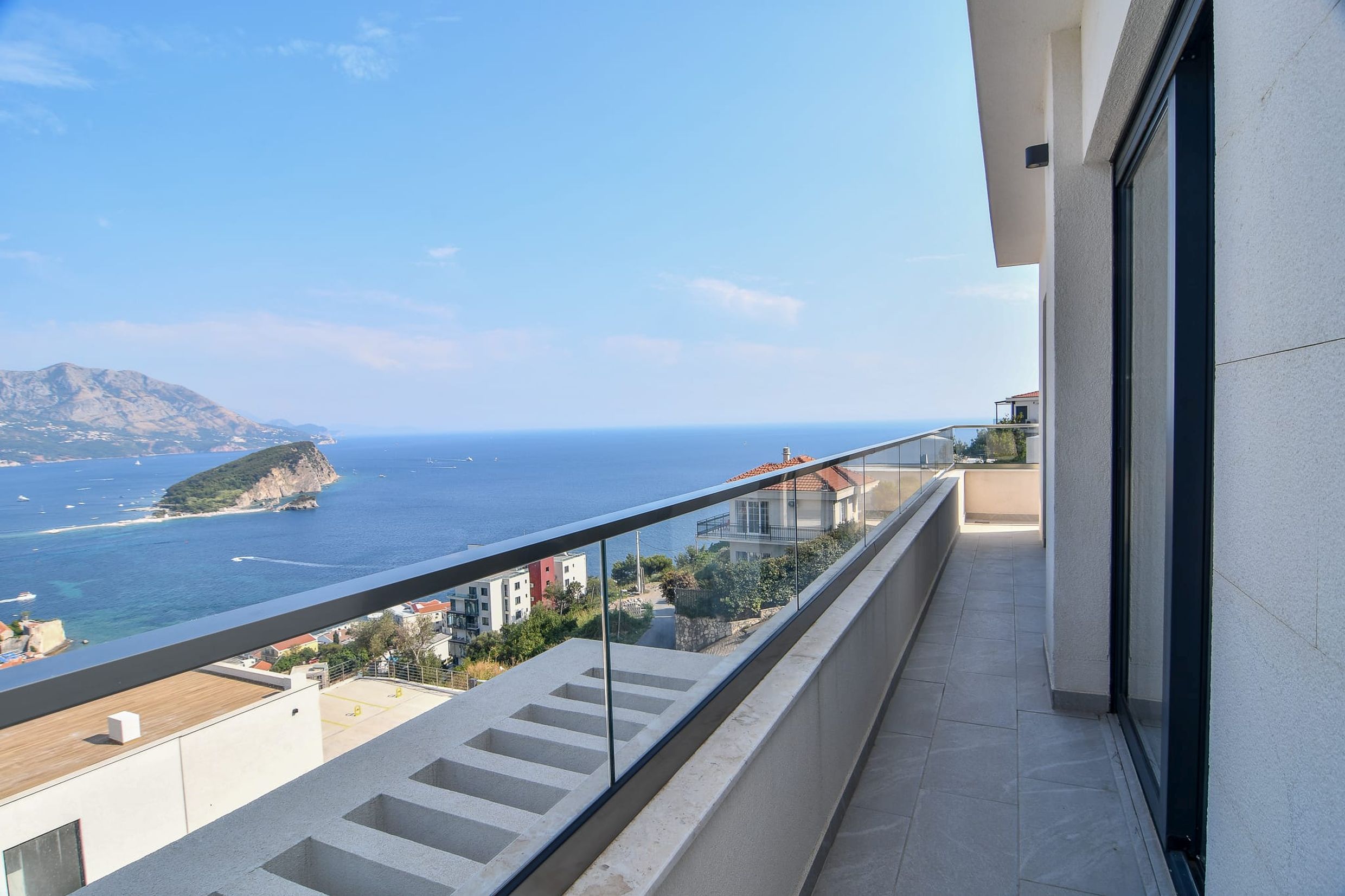 Spacious balcony with panoramic views of the sea and island in Budva, Montenegro: modern finishes and glass railings.