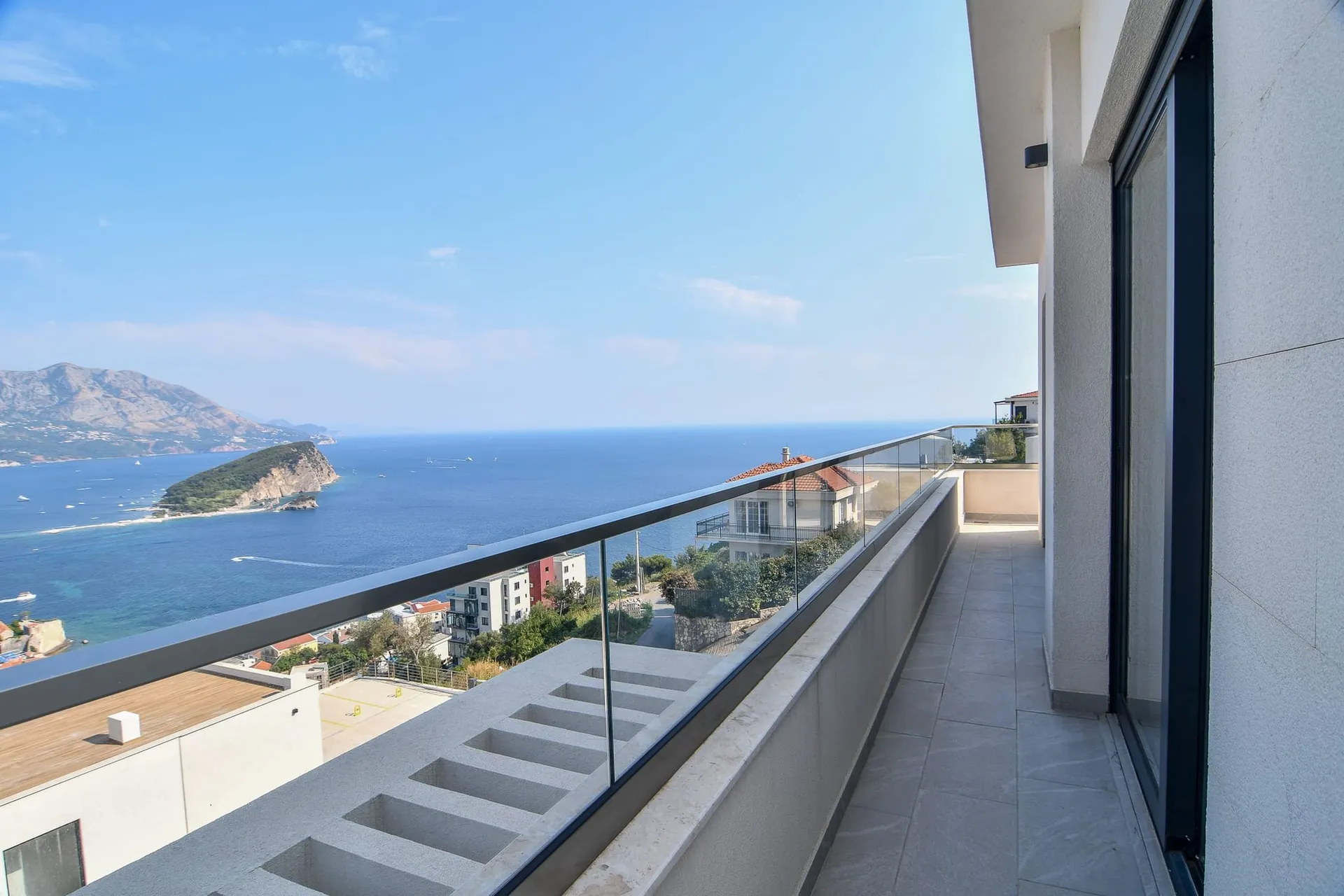 Wide balcony with glass railing offering a panoramic view of the sea, island, and mountains.