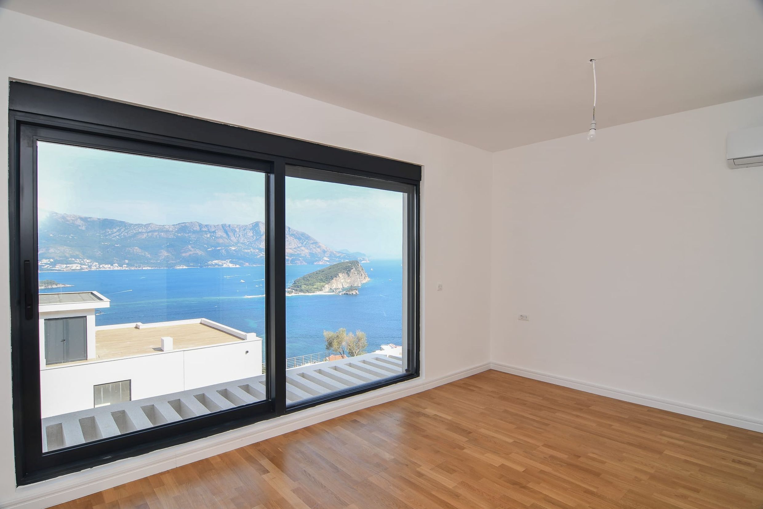 Bright living room with panoramic view of the sea and island in Budva, Montenegro: parquet floor, large windows, air conditioner.