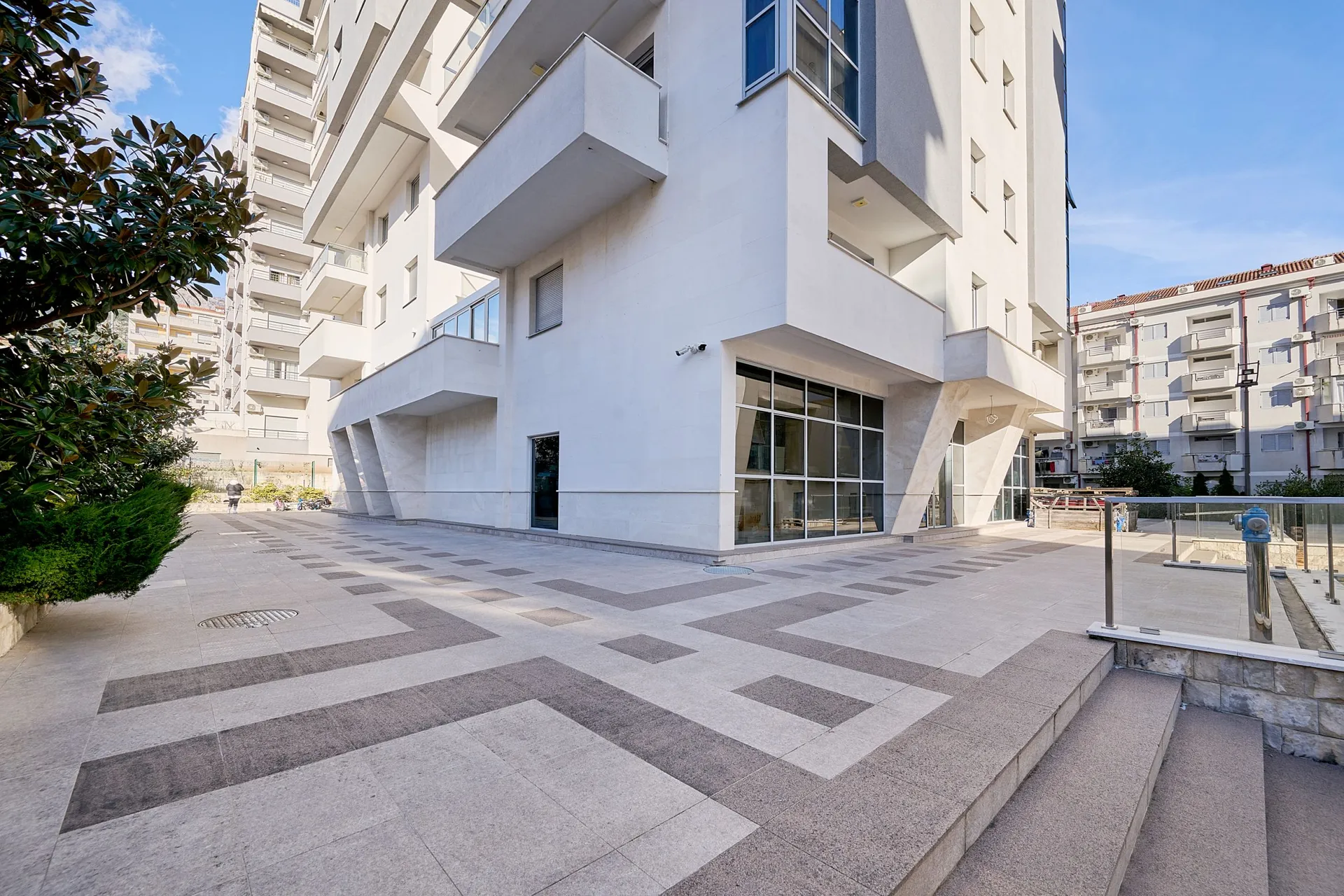 Exterior view of a modern white residential building with balconies and a geometric pattern on the stone pavement.