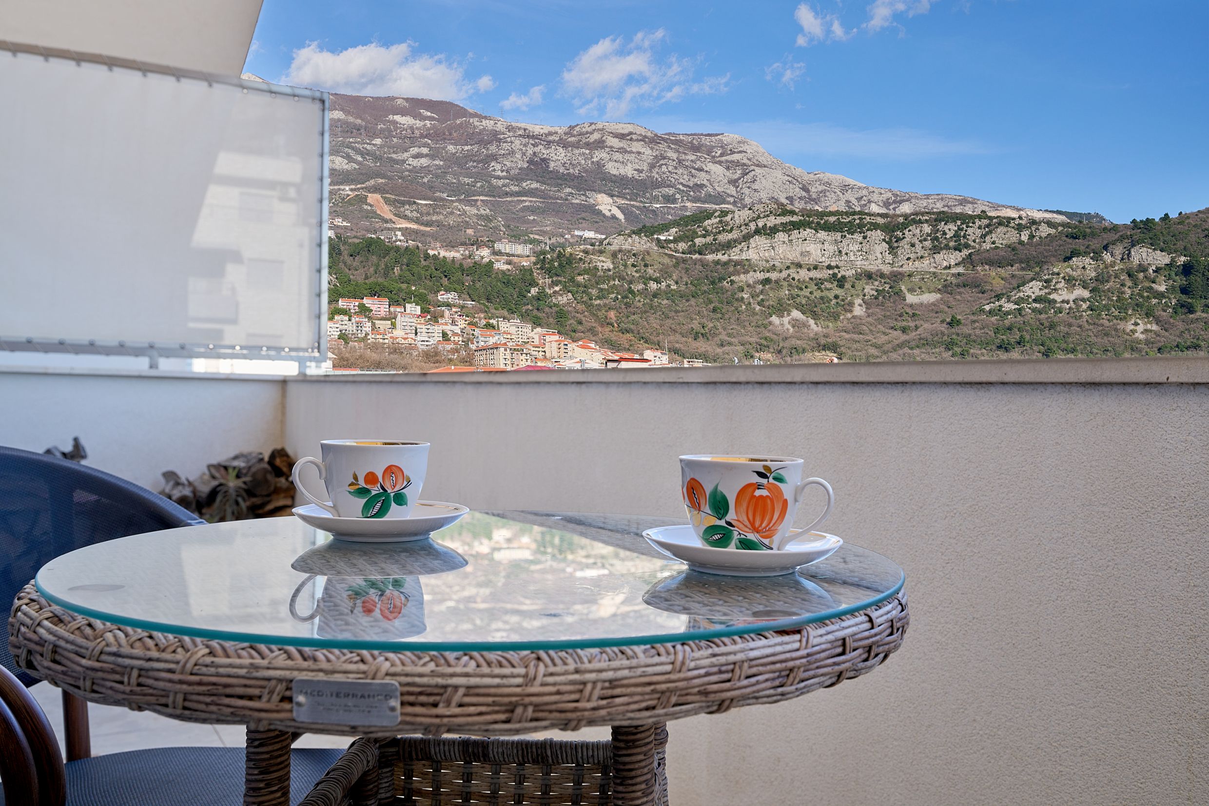 View from the apartment balcony in Budva, Montenegro: a wicker table with cups against a mountain and city backdrop.