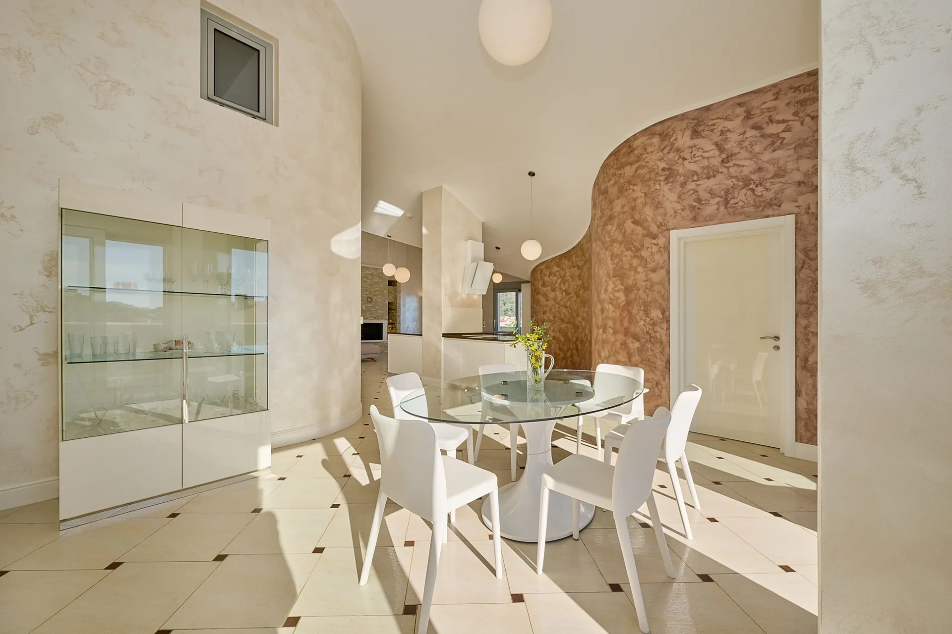 Bright kitchen with white chairs, a glass table, and a display cabinet. An arched wall with marble texture is visible.