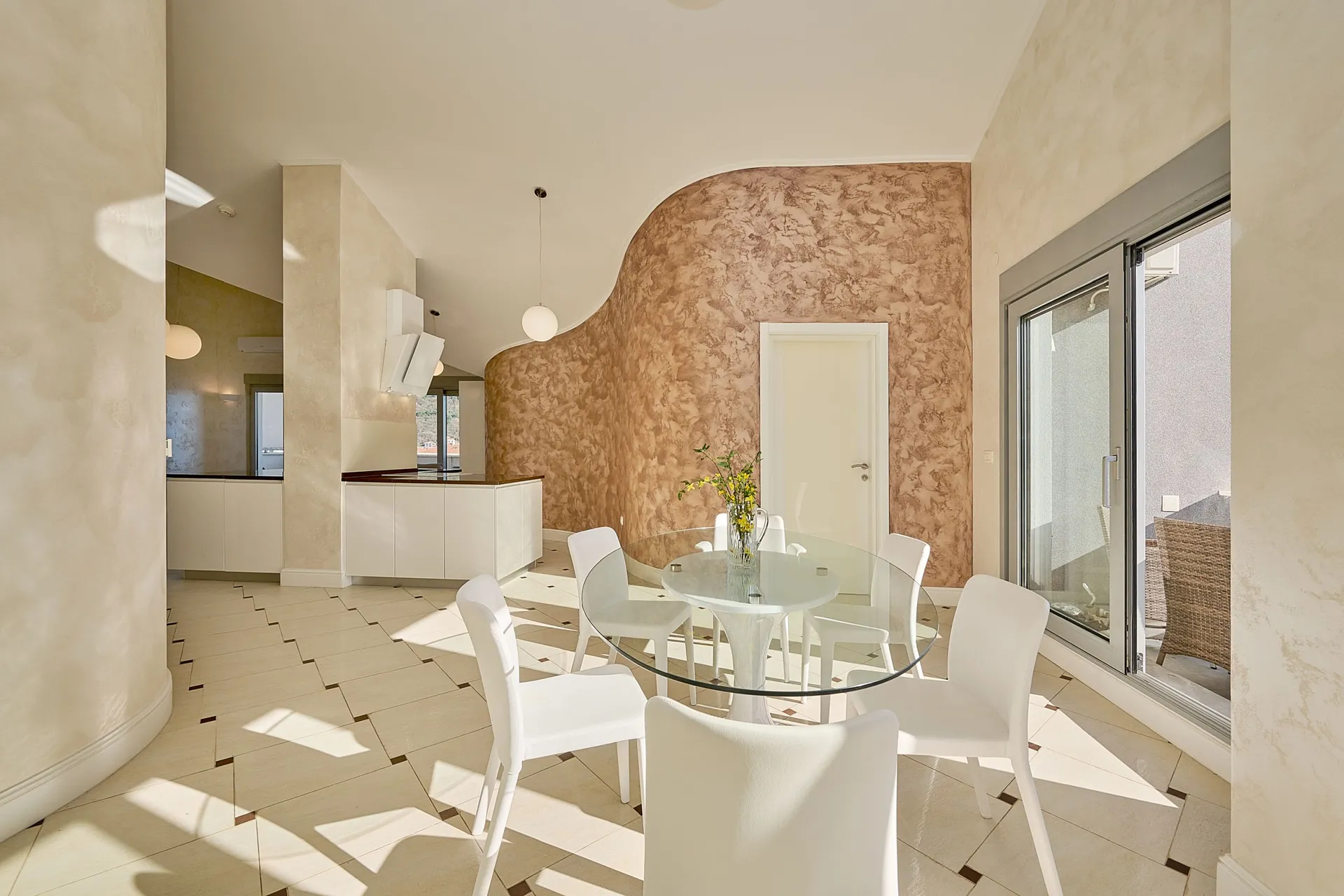 Bright kitchen-dining area with white chairs and a round glass table. The background shows a kitchen zone with white furniture and a decorative wall.