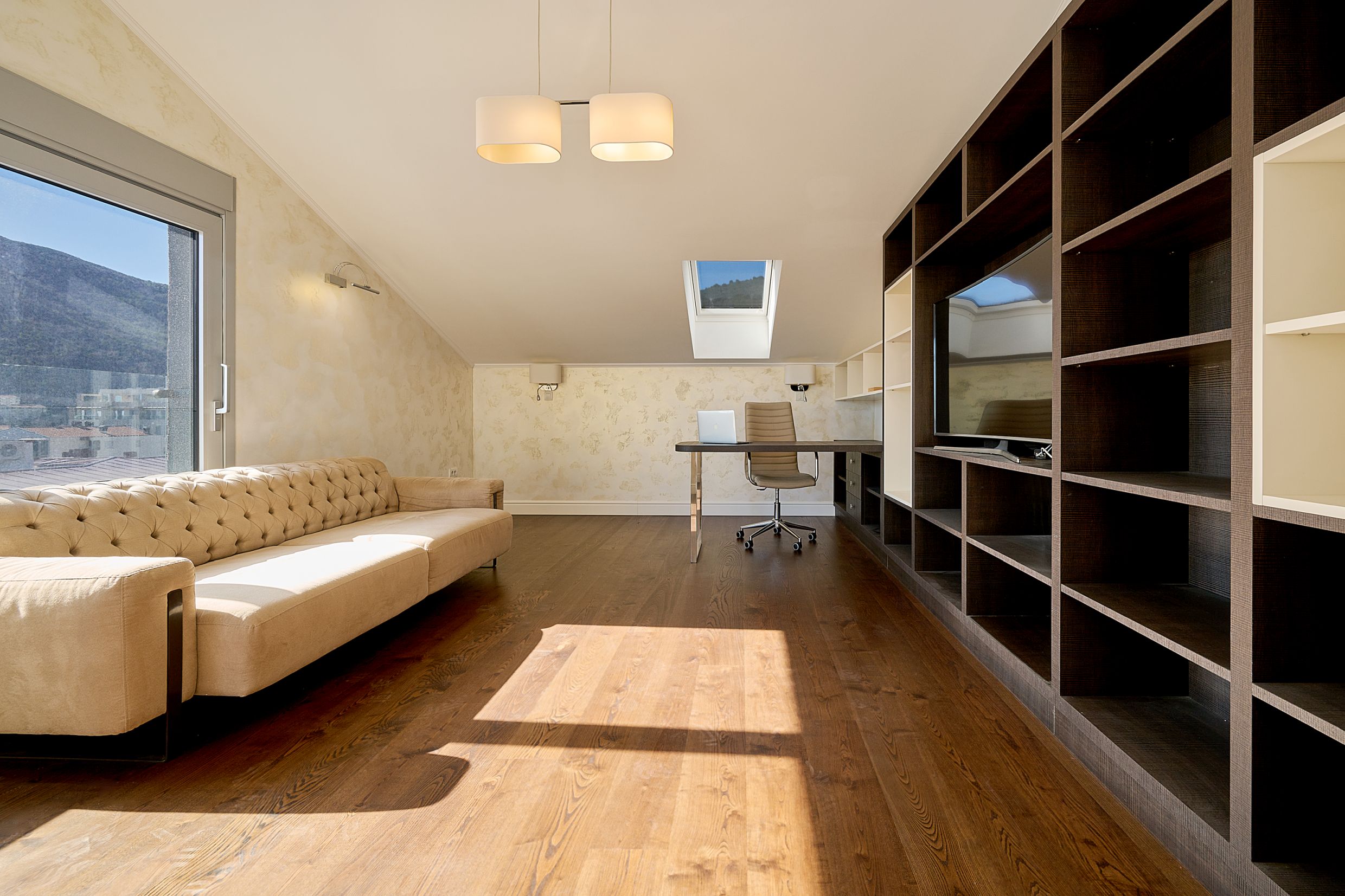 Modern living room with a home office in an apartment in Budva, Montenegro: beige sofa, dark shelving unit, and mountain view.