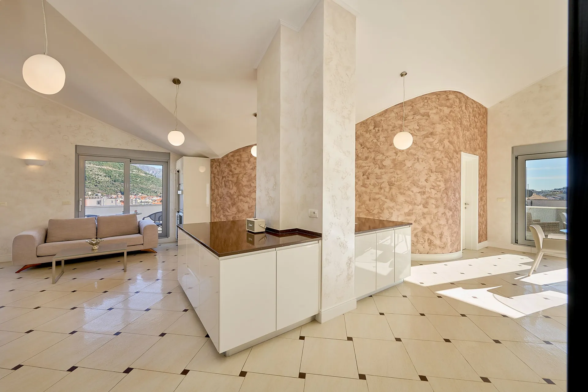 Bright living room with a beige sofa, white kitchen island, decorative textured walls, and tiled flooring.