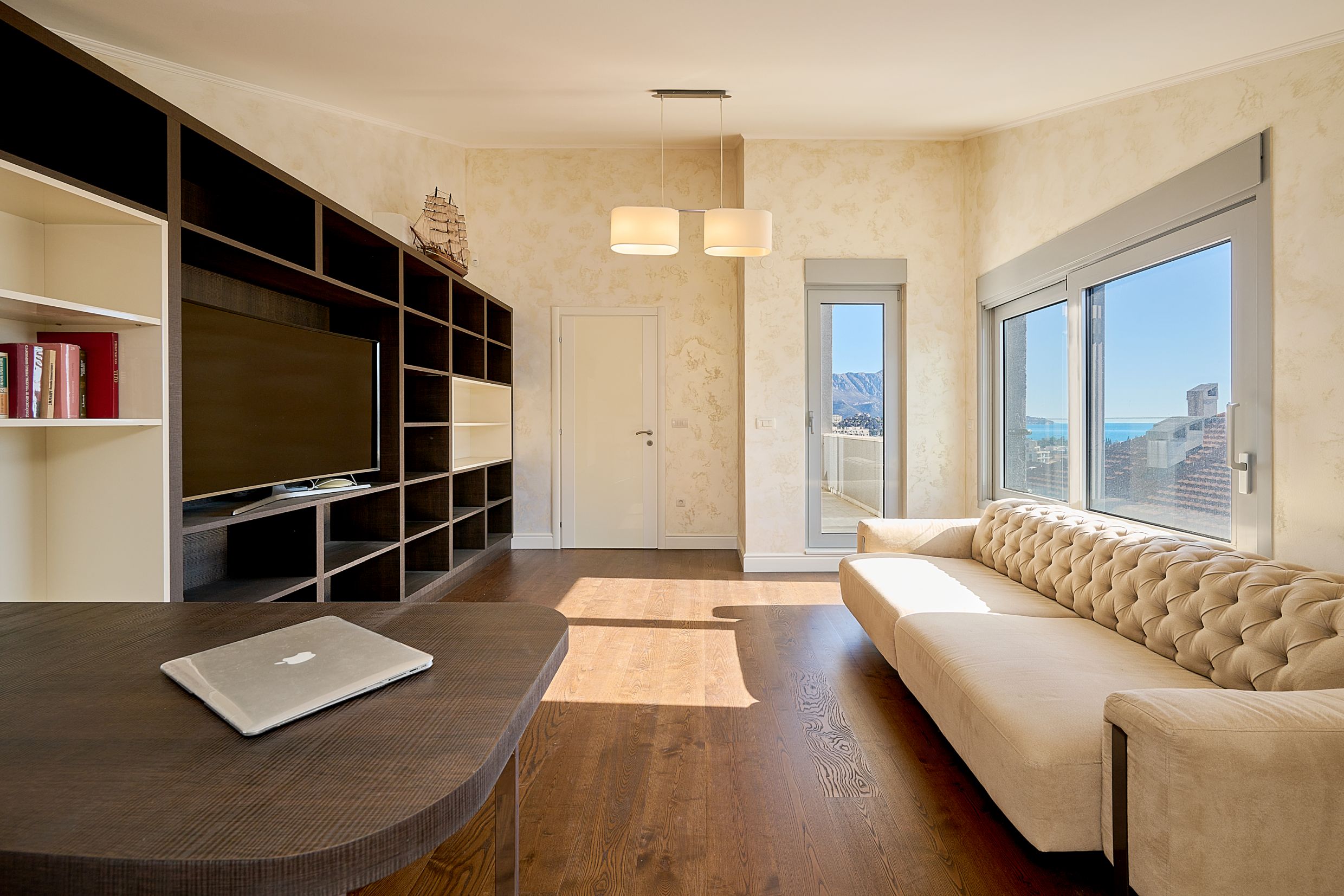 Living room in an apartment in Budva, Montenegro: a light sofa, dark shelving unit, parquet flooring, and a panoramic view of the sea and mountains.