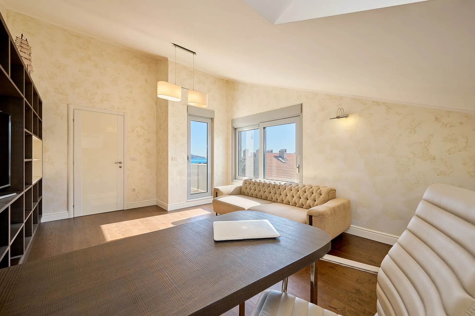Bright room with a beige sofa, dark table, and shelving unit. A laptop sits on the table near windows overlooking the city.