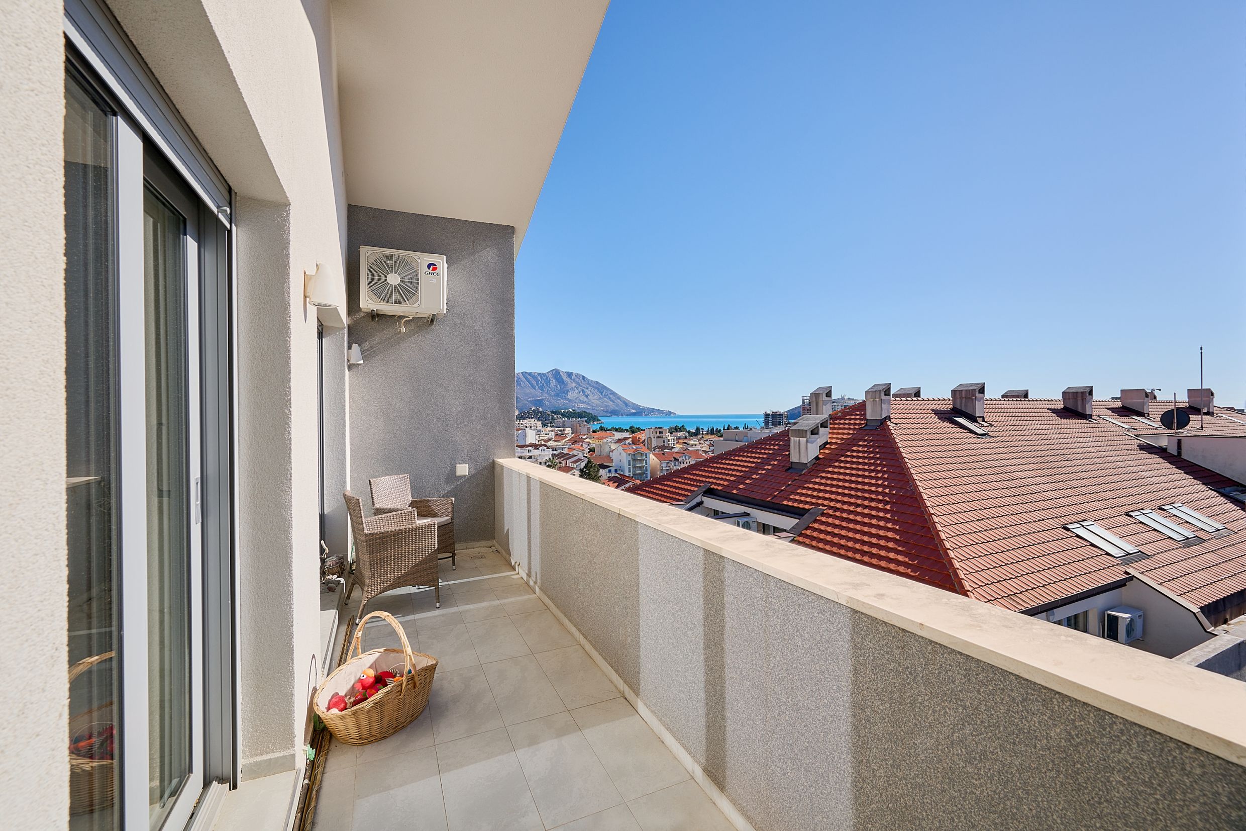 Apartment balcony in Budva, Montenegro: wicker furniture, sea view, mountains, and the city’s tiled roofs.
