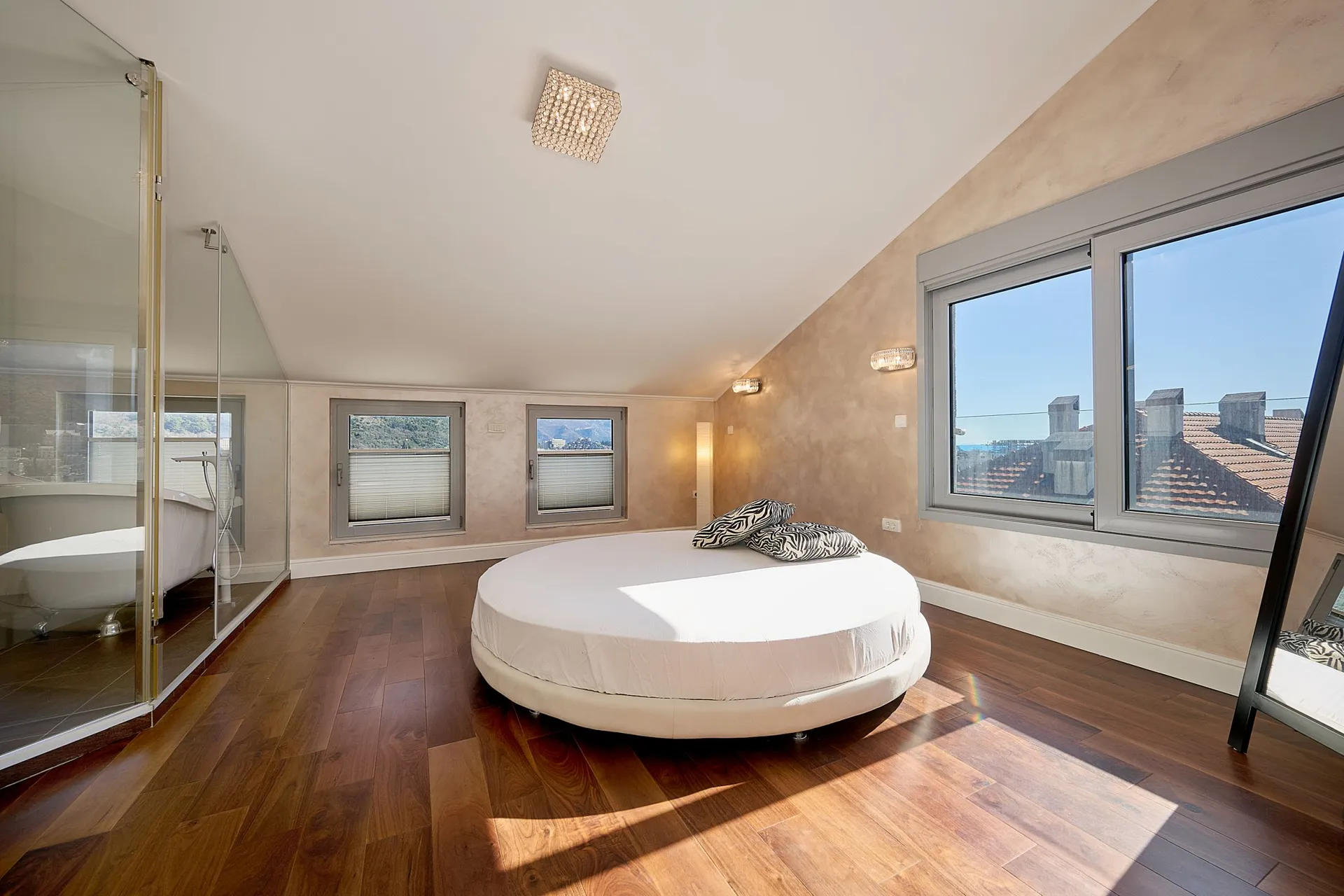 Modern bedroom with a round bed and wooden floor. A glass partition on the left reveals an adjacent bathtub area.