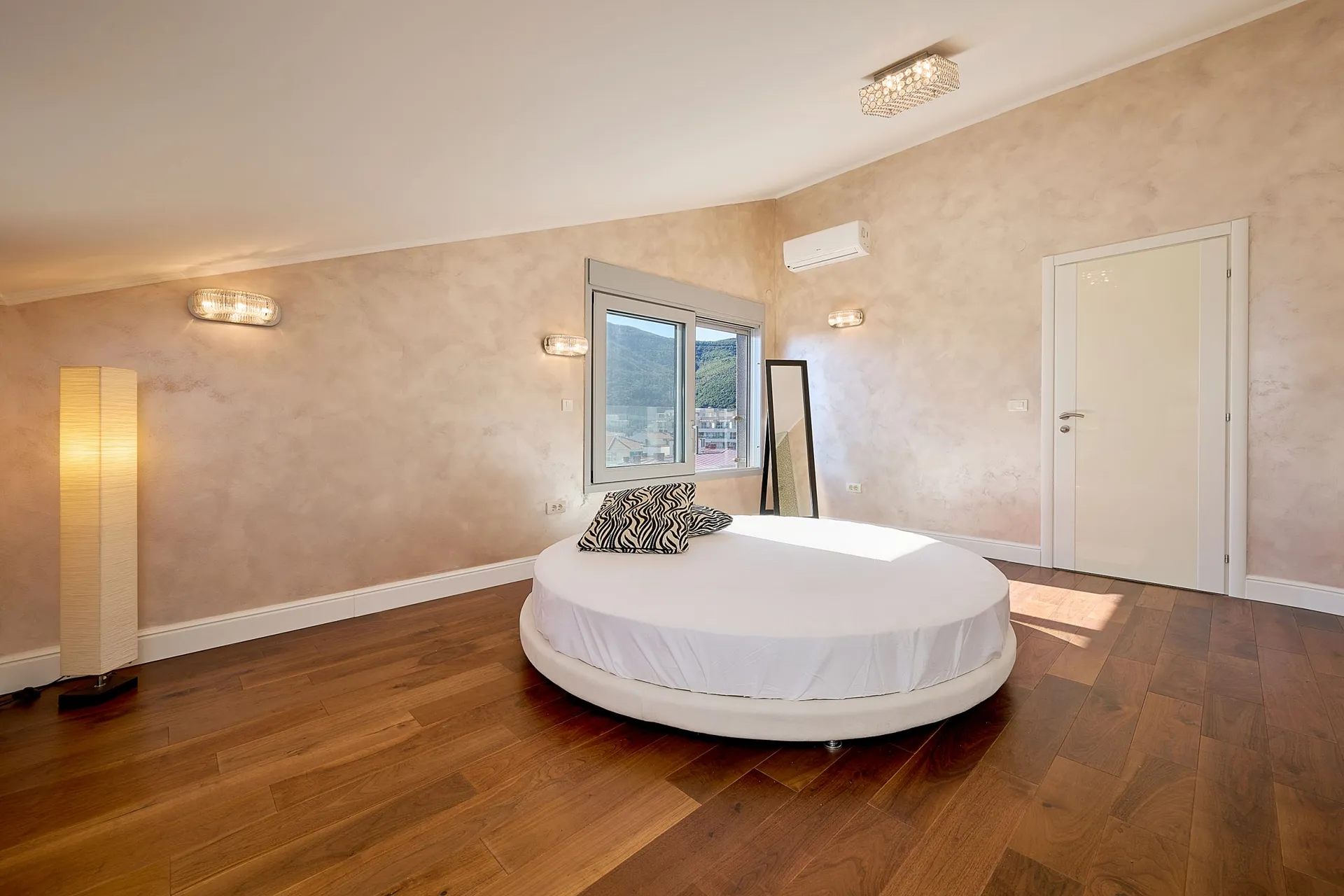 Modern bedroom featuring a round white bed, dark wood flooring, and textured beige walls.