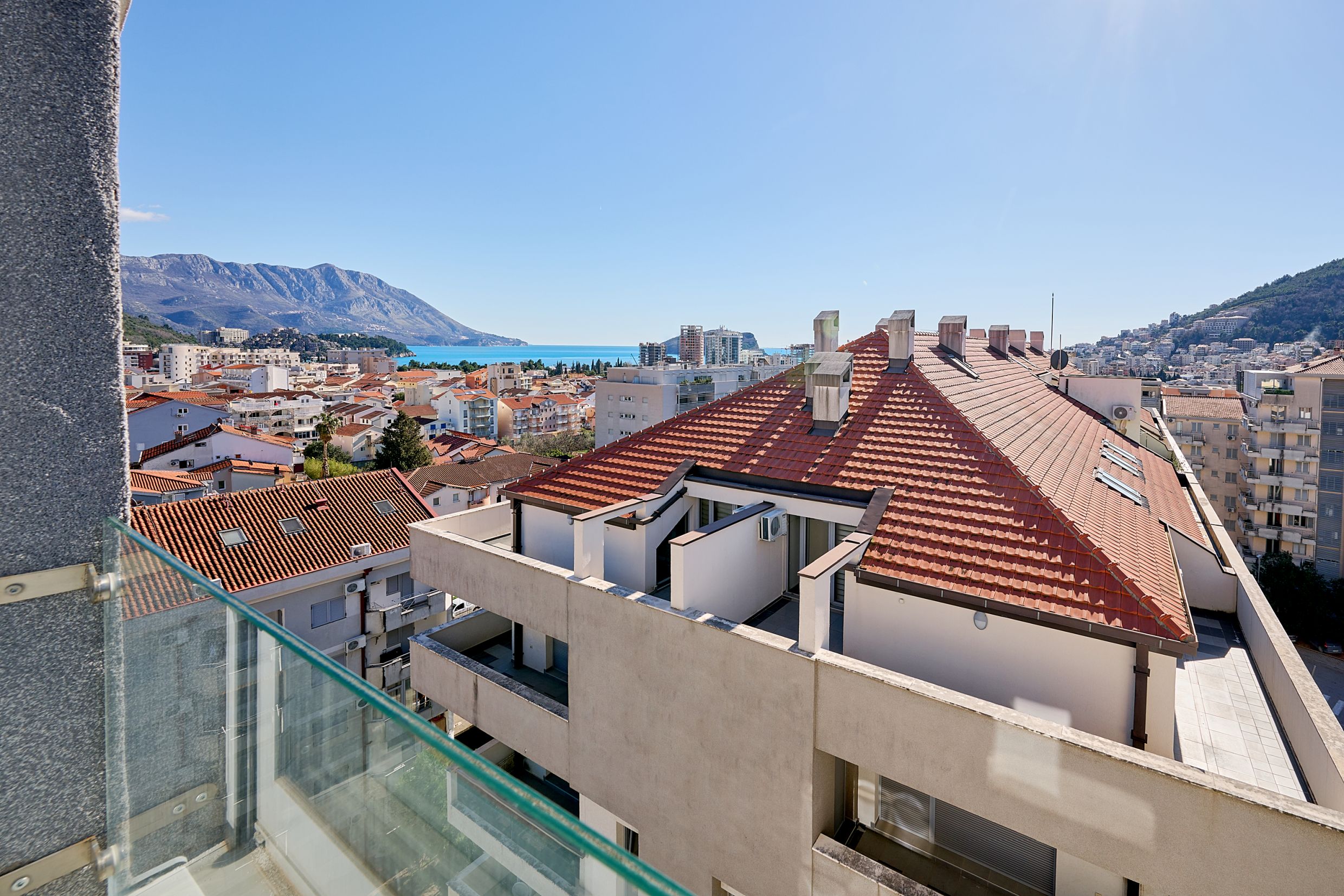 View from the apartment balcony in Budva, Montenegro: city panorama, tiled roofs, mountains, and the Adriatic Sea.