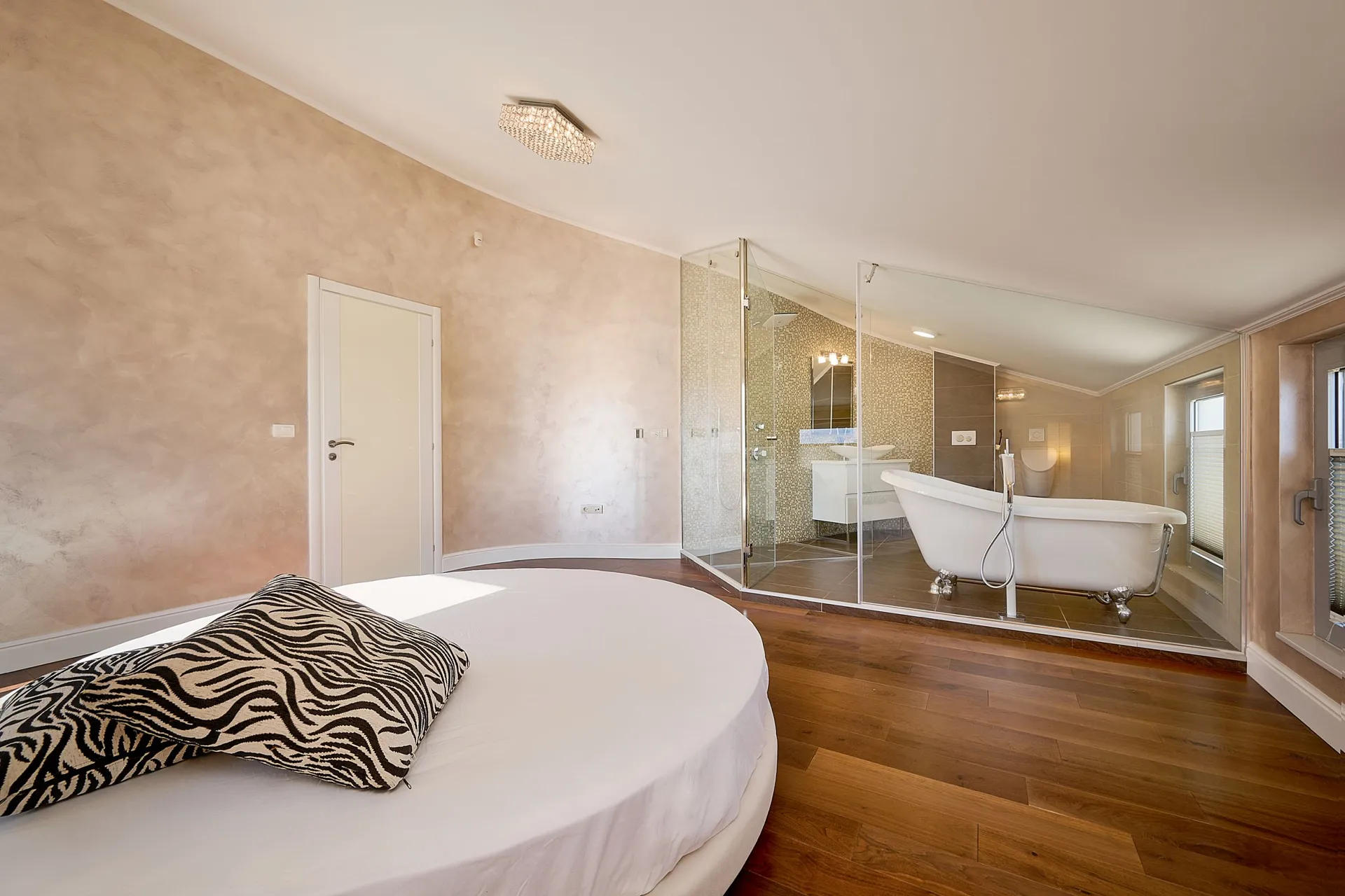 Bedroom with a round bed and decorative pillow. An adjacent bathroom is visible behind a glass partition featuring a freestanding bathtub.