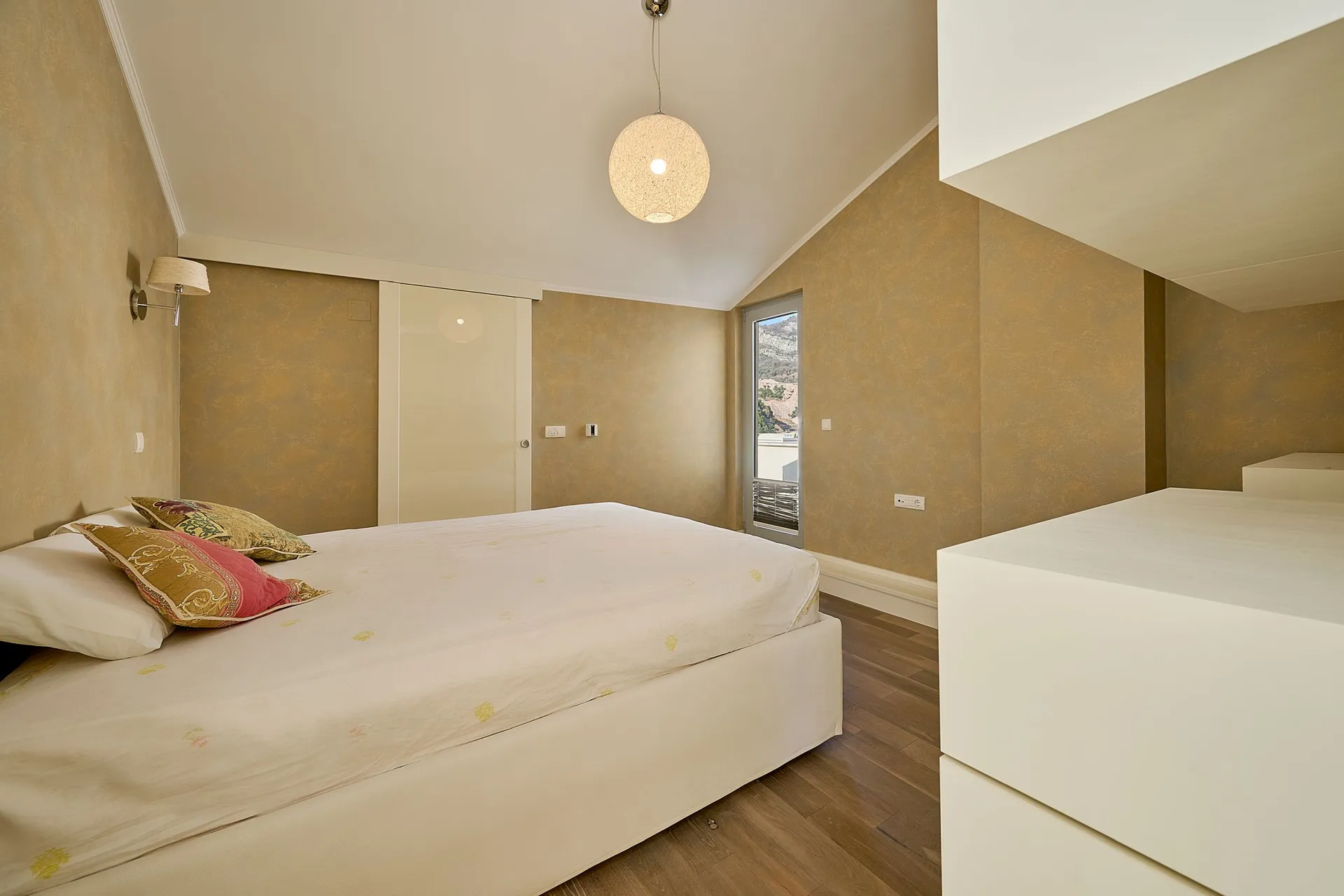 Modern bedroom featuring a white bed, golden textured walls, a round pendant light, and dark wooden flooring.