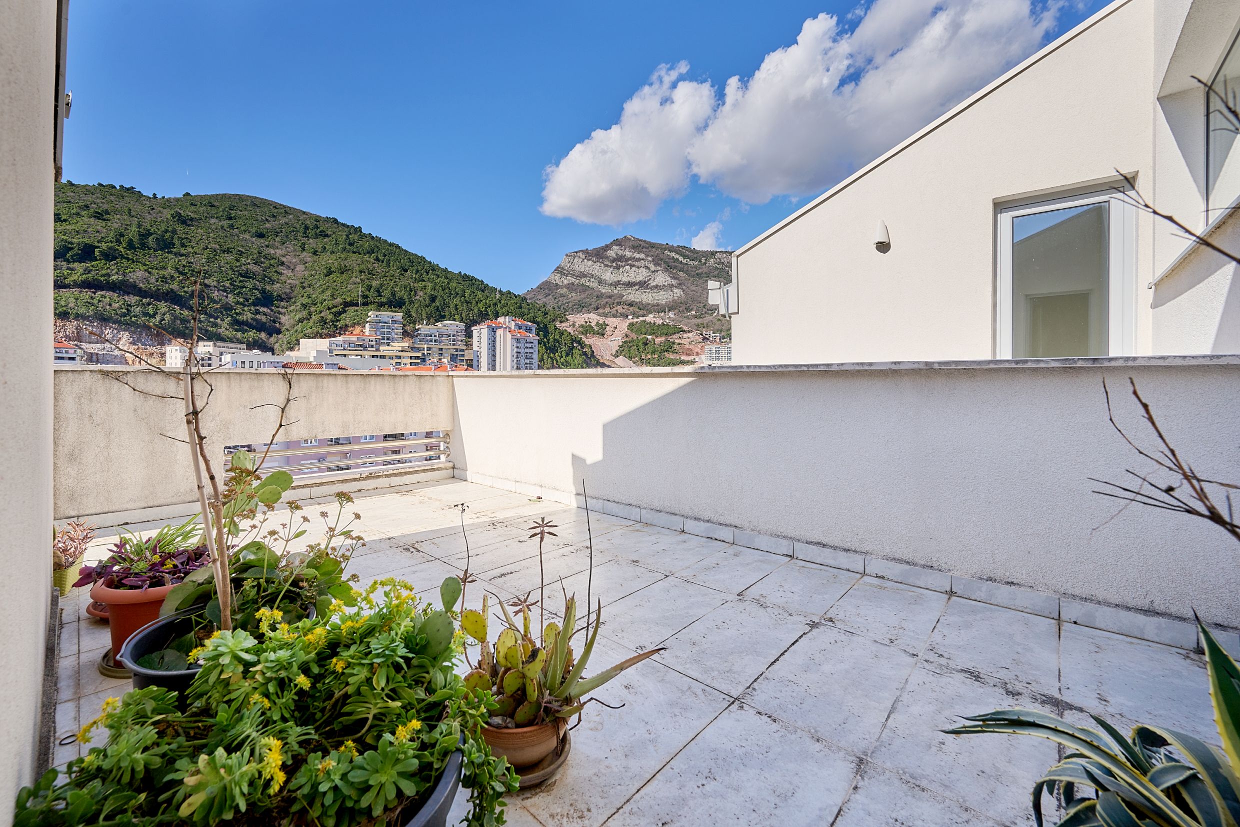 Spacious terrace with plants and mountain views in an apartment in Budva, Montenegro. Light tile and an open balcony.