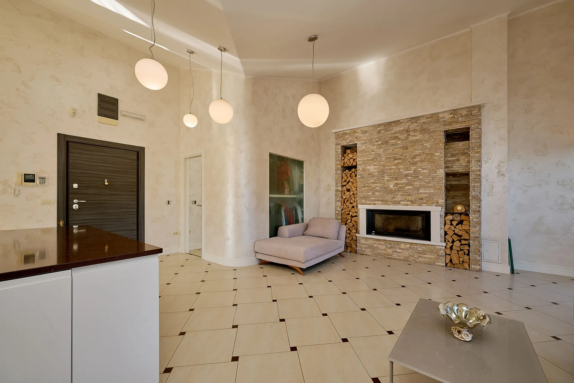 Bright room featuring a stone fireplace with wood storage shelves, spherical pendant lights, a grey chaise lounge, and tiled flooring.