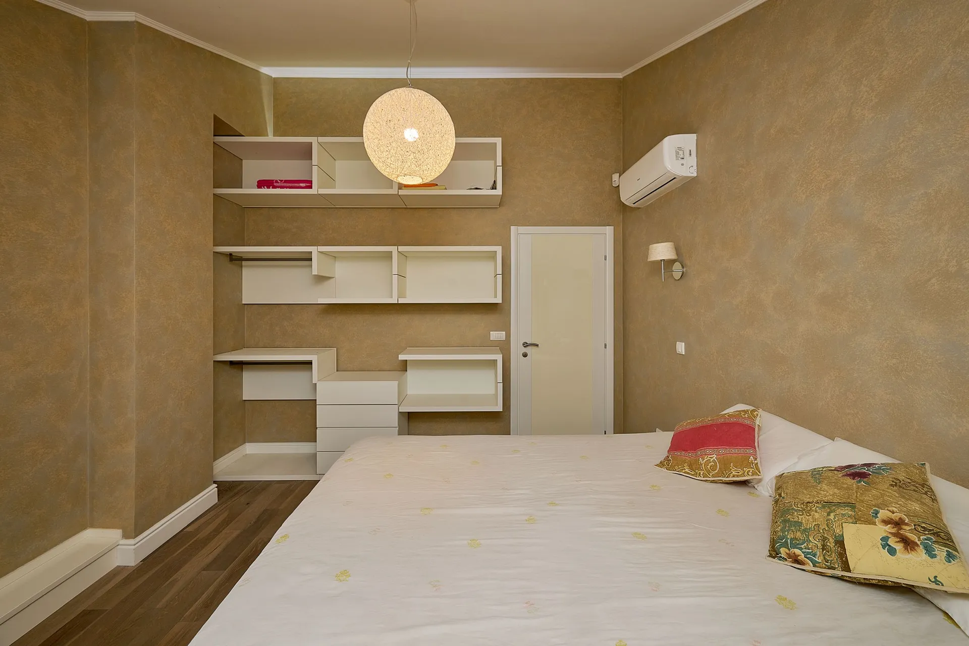 Modern bedroom featuring a white bed, built-in shelving units, and a round pendant light against textured beige walls.