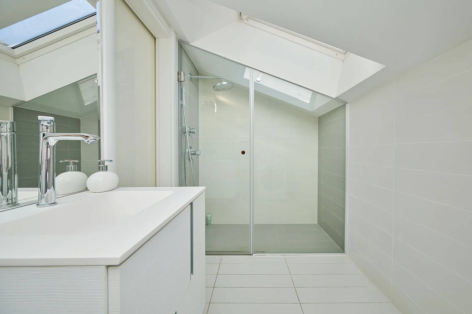 Modern white bathroom featuring a glass shower enclosure under a skylight and a vanity with a chrome faucet.
