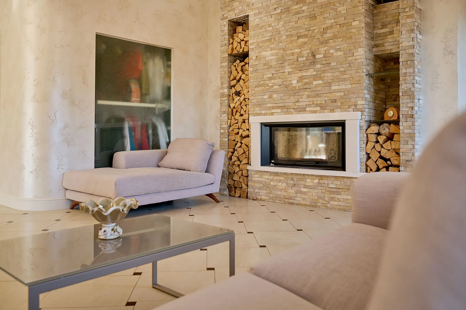 Bright living room featuring a stone fireplace with stacked firewood, a beige chaise lounge, and a glass coffee table on tiled flooring.