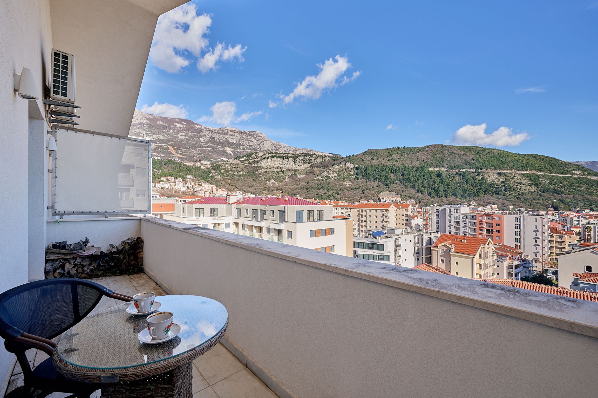 View from the apartment balcony in Budva, Montenegro: a table with cups, city panorama, and mountain scenery on a sunny day.