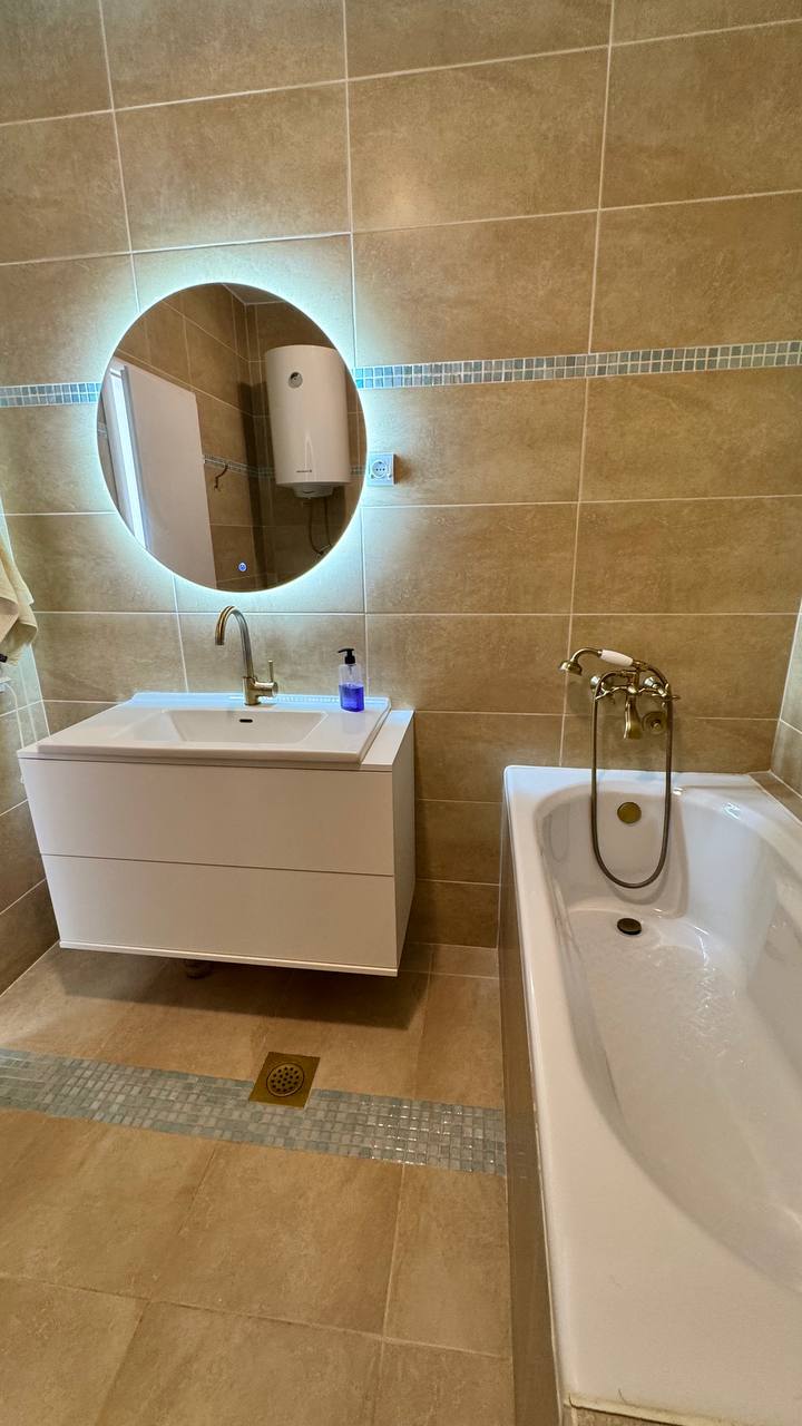 Bathroom in an apartment in Budva, Montenegro: a white vanity with sink, a round backlit mirror, and a bathtub.