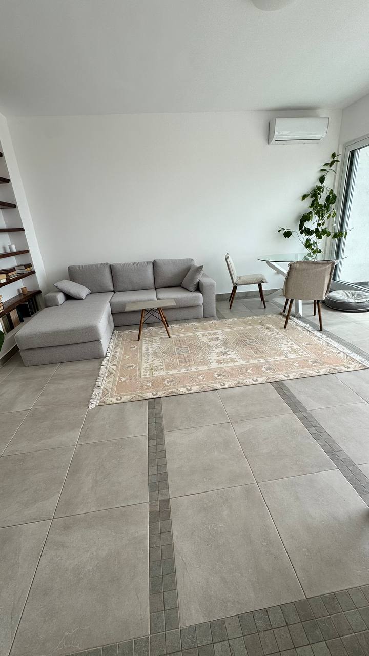 Bright living room in an apartment in Budva, Montenegro: gray corner sofa, patterned rug, dining area, and panoramic window.