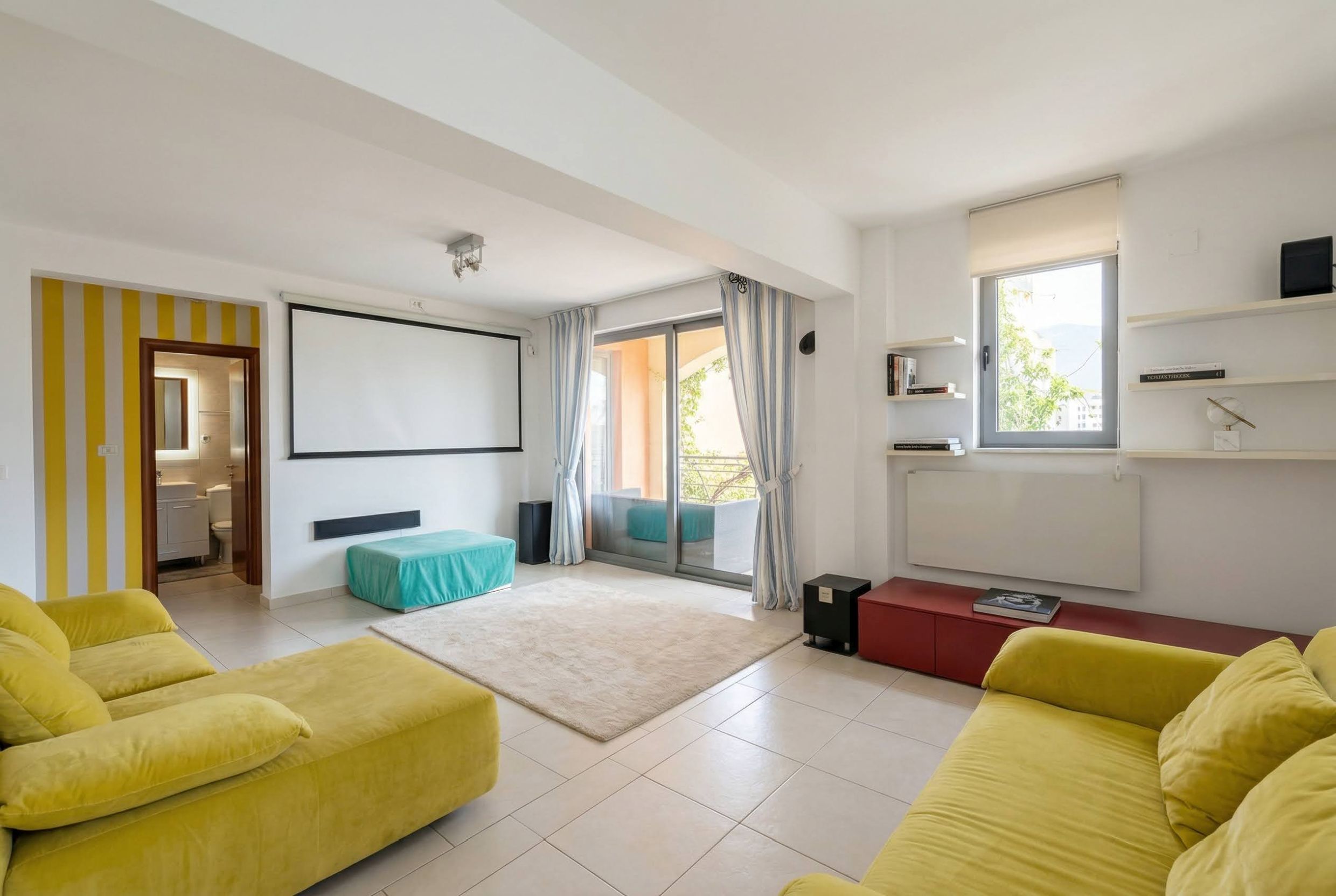 Bright living room with a projector and yellow sofas in an apartment in Bečići, Montenegro. Access to the terrace and garden view.