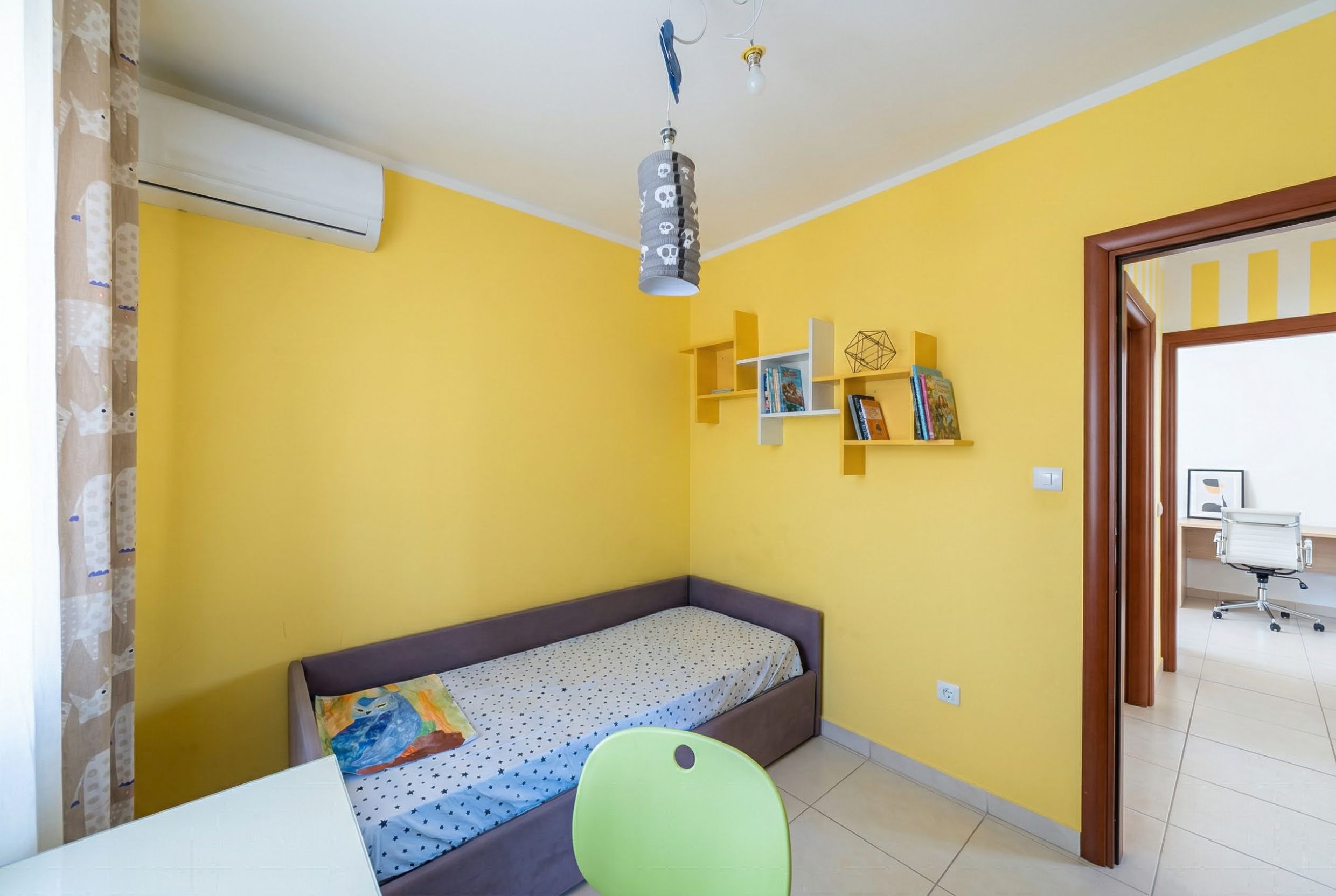Children’s bedroom with yellow walls and a bed in an apartment, Becici, Montenegro. Cozy interior with shelves and air conditioning.