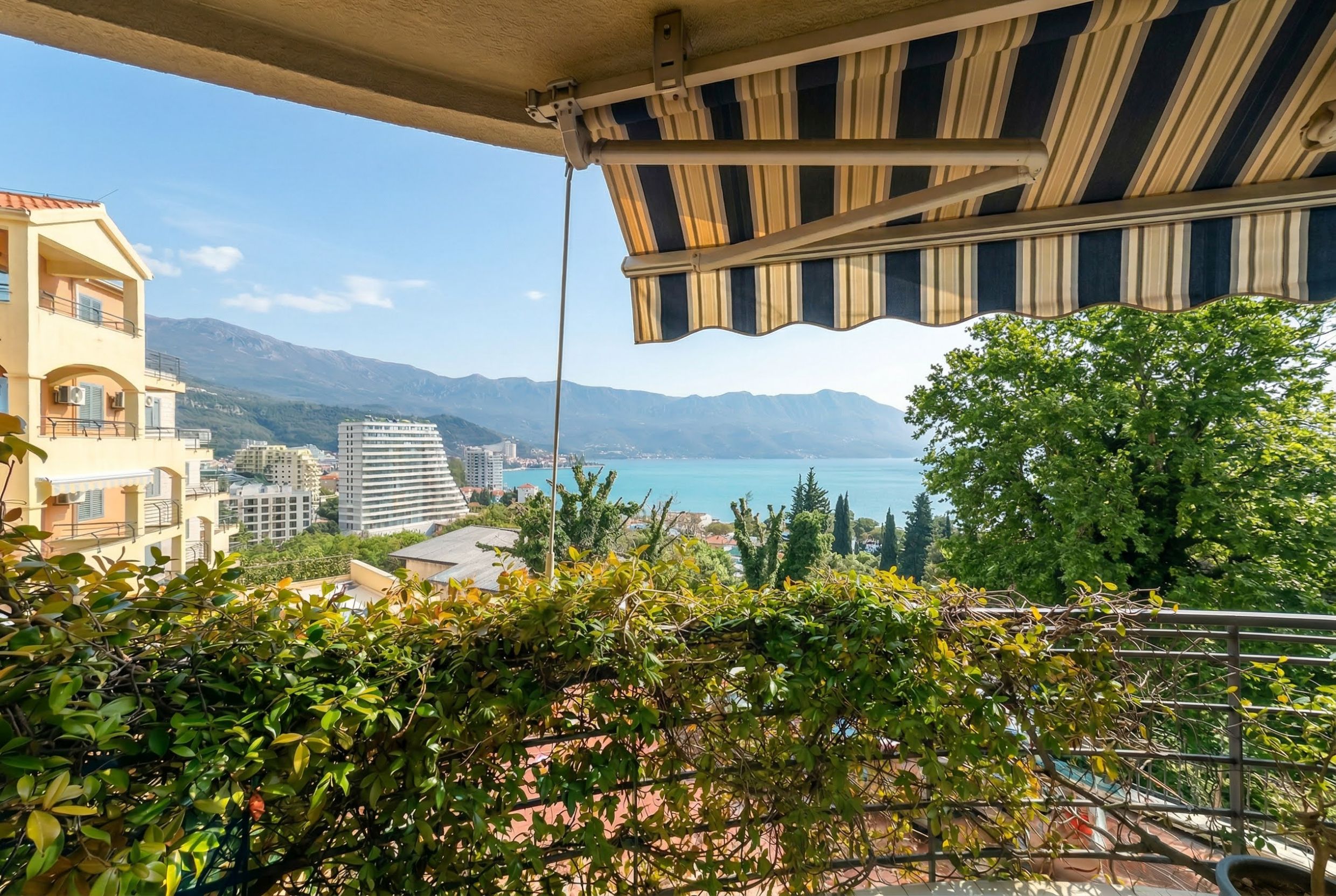 View from the apartment balcony in Bečići, Montenegro: a panorama of the sea, mountains, and a striped sun awning over the terrace.