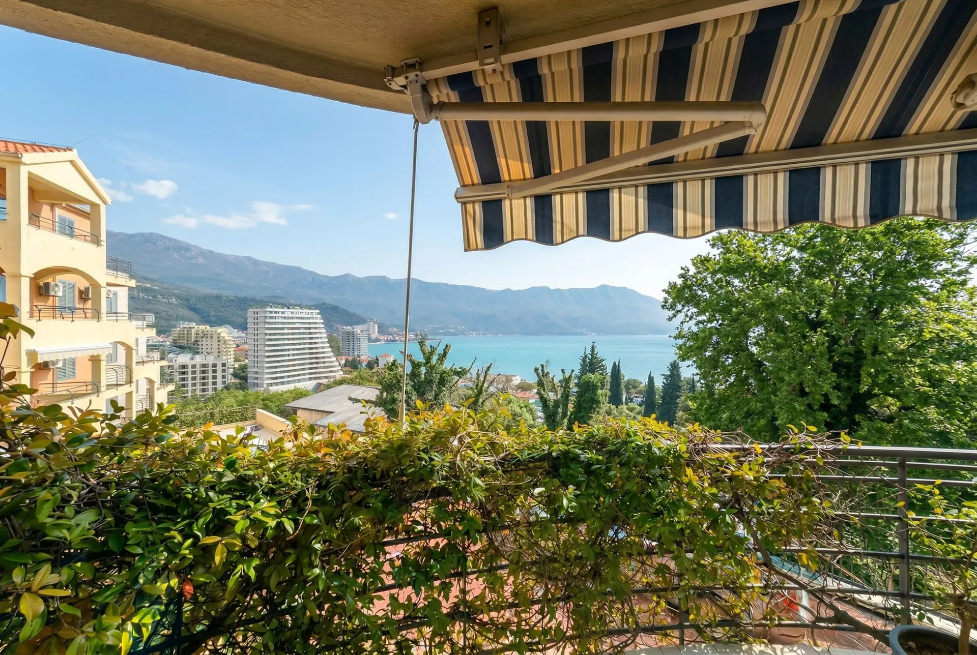 View from a balcony over the sea and mountains. A striped awning is above, with dense greenery and a metal railing in front.