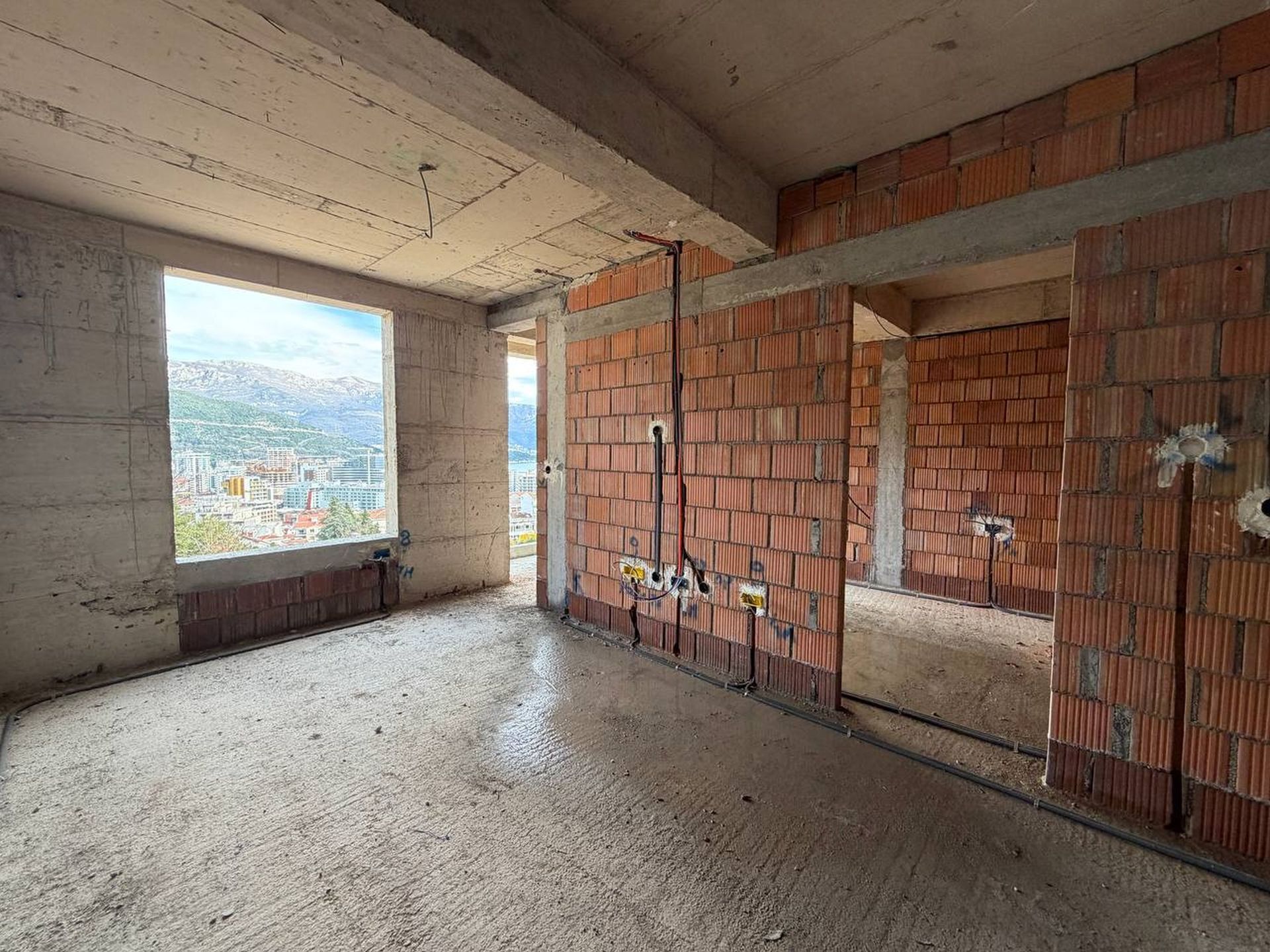 Unfinished living room in a new building in Budva, Montenegro: brick walls, concrete floor, and panoramic city view.