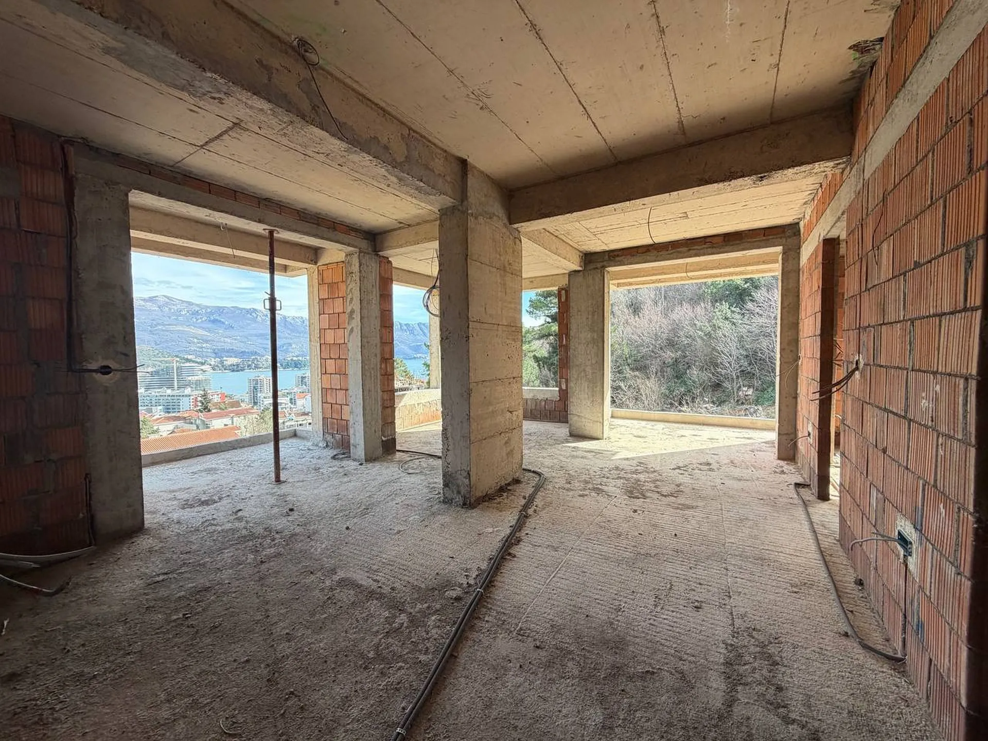 Unfinished construction space with concrete columns, brick walls, and rough floor overlooking a city and sea.