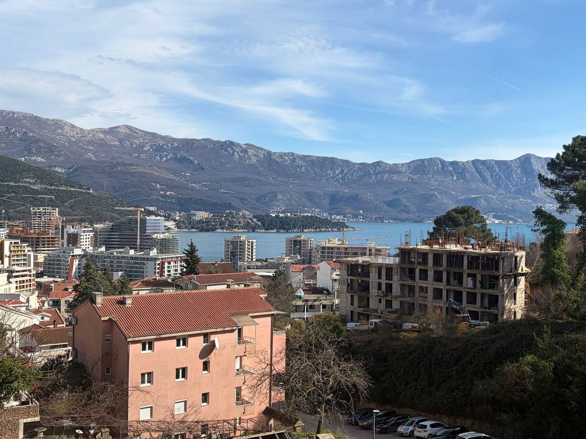 Panoramic view of Budva, Montenegro: residential buildings, construction sites, and the sea bay against a backdrop of mountain peaks.