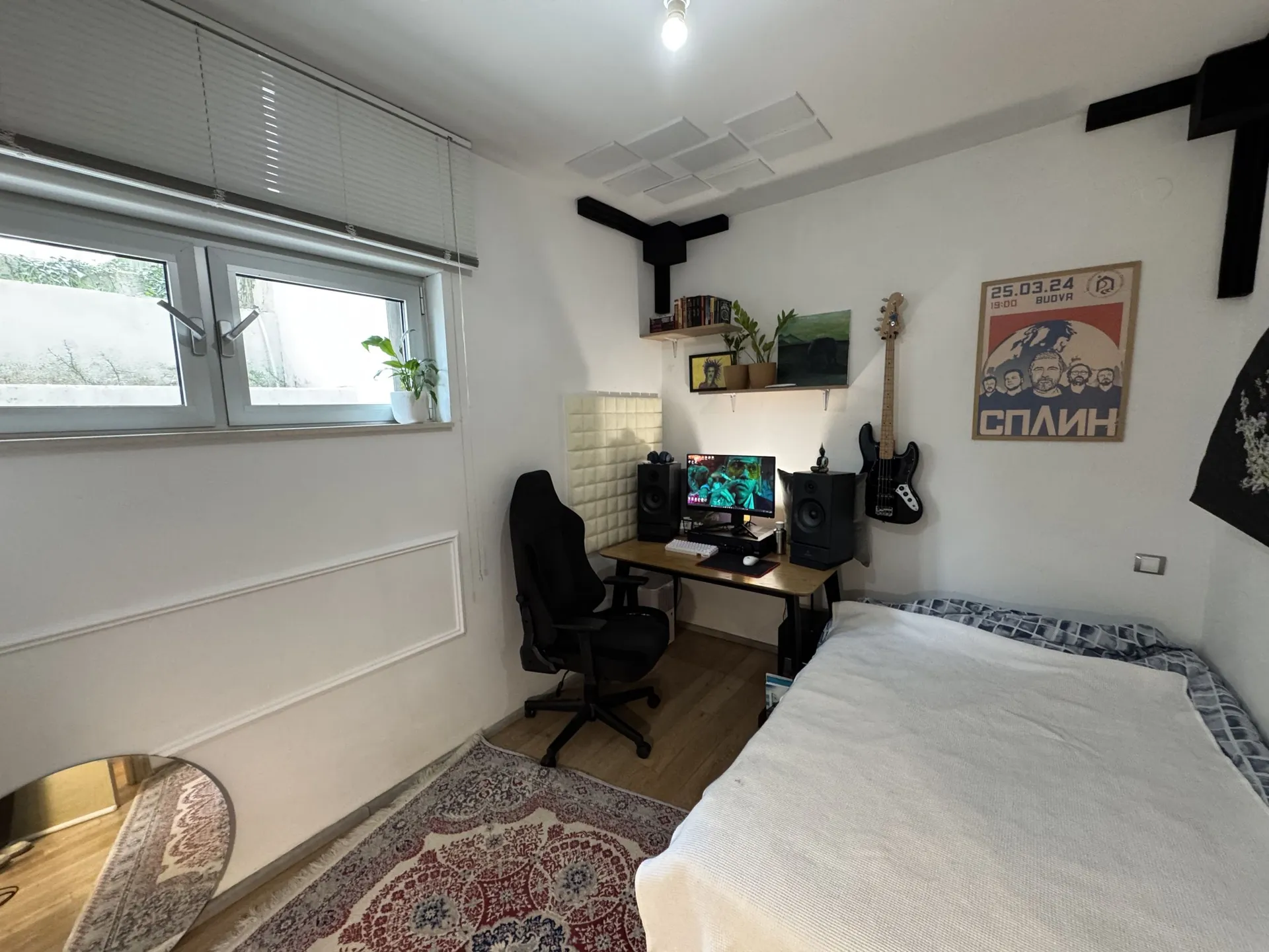 Bedroom with a computer desk, gaming chair, bass guitar on the wall, and a poster. Part of the bed and rug are visible.