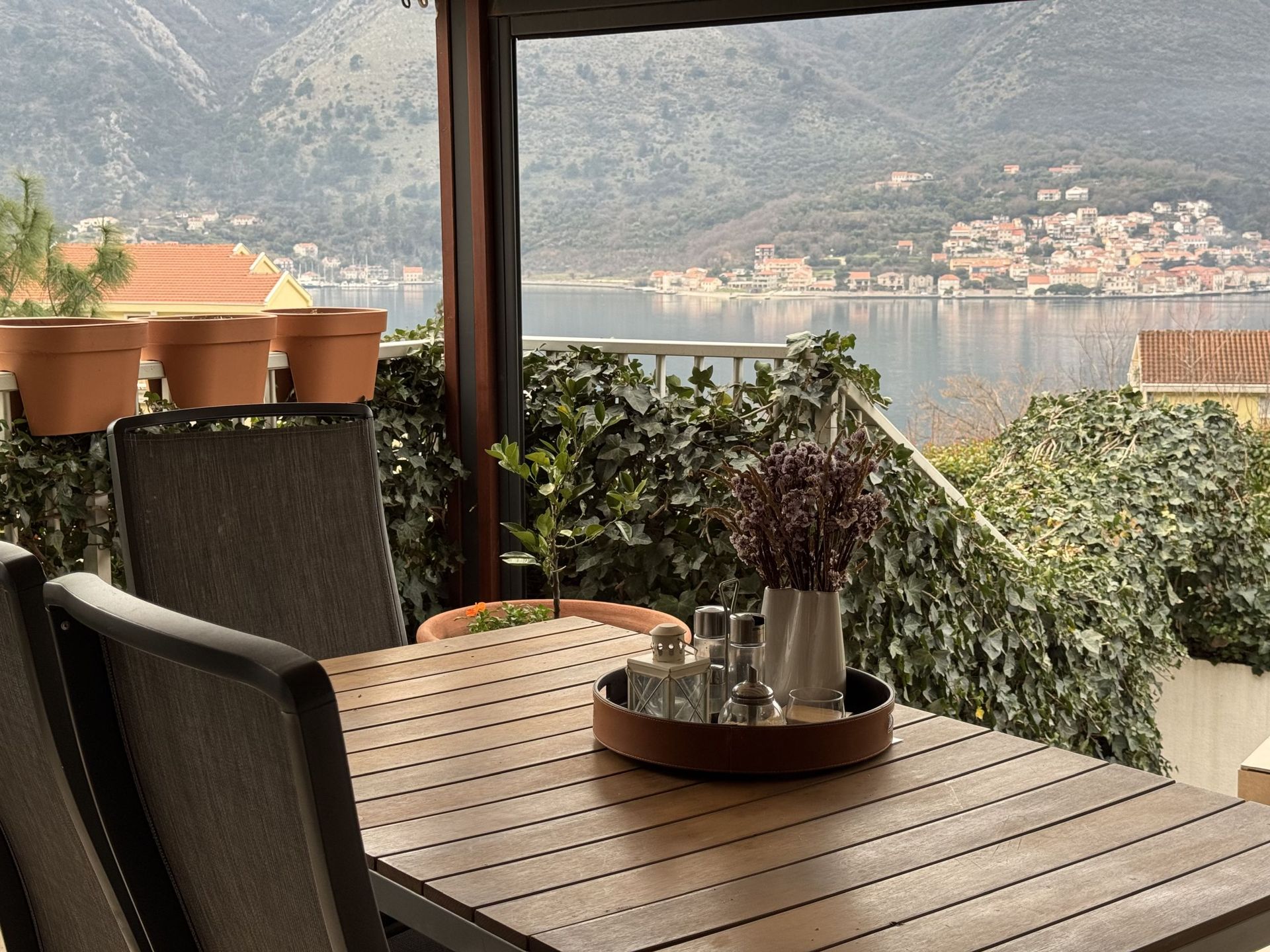 Terrace with a wooden table and a view of Kotor Bay in Dobrota, Montenegro. Cozy balcony with plants and decor.