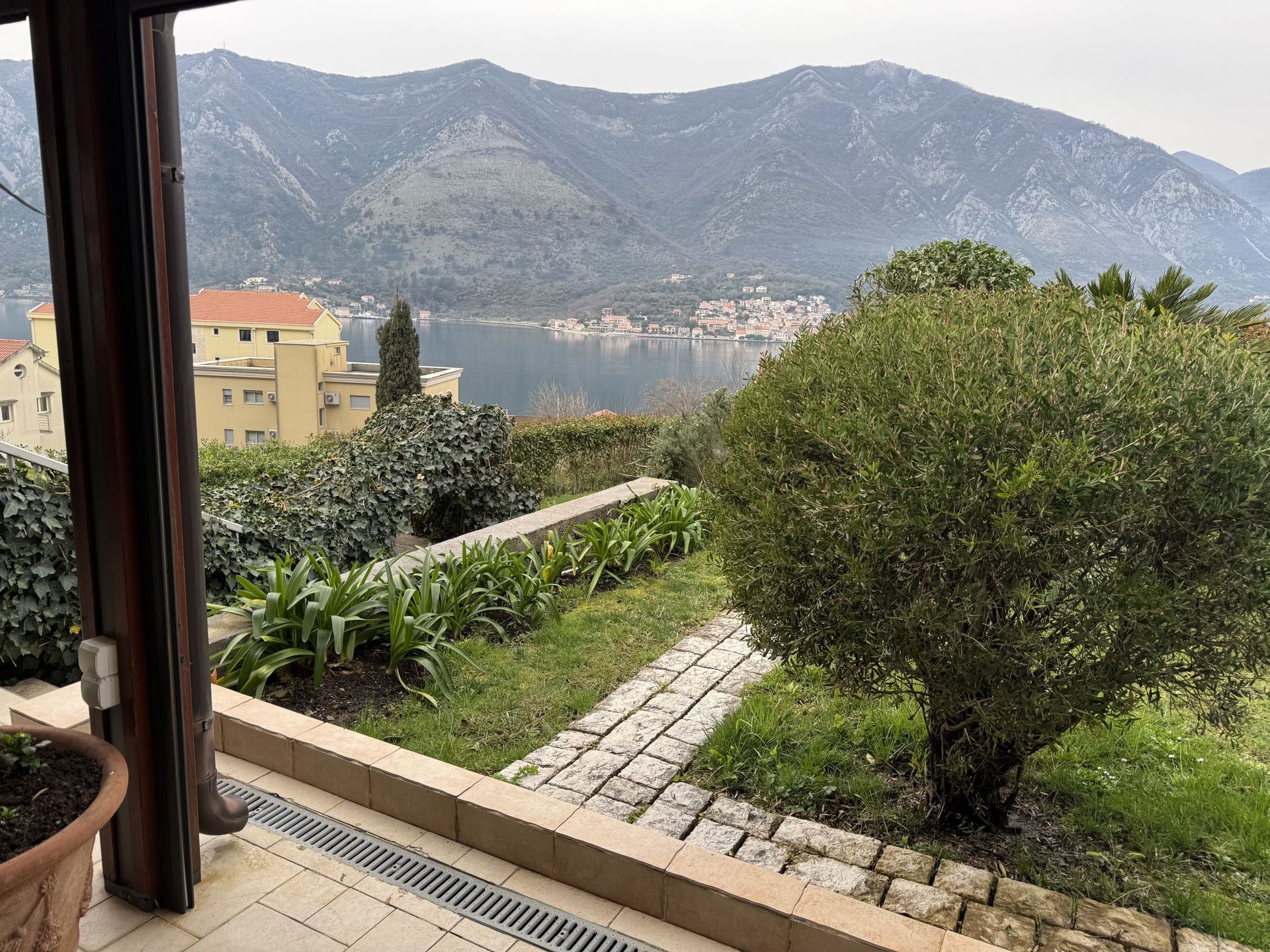 View from the apartment terrace in Dobrota, Montenegro: a garden with a stone path, panoramic view of the Bay of Kotor and the mountains.