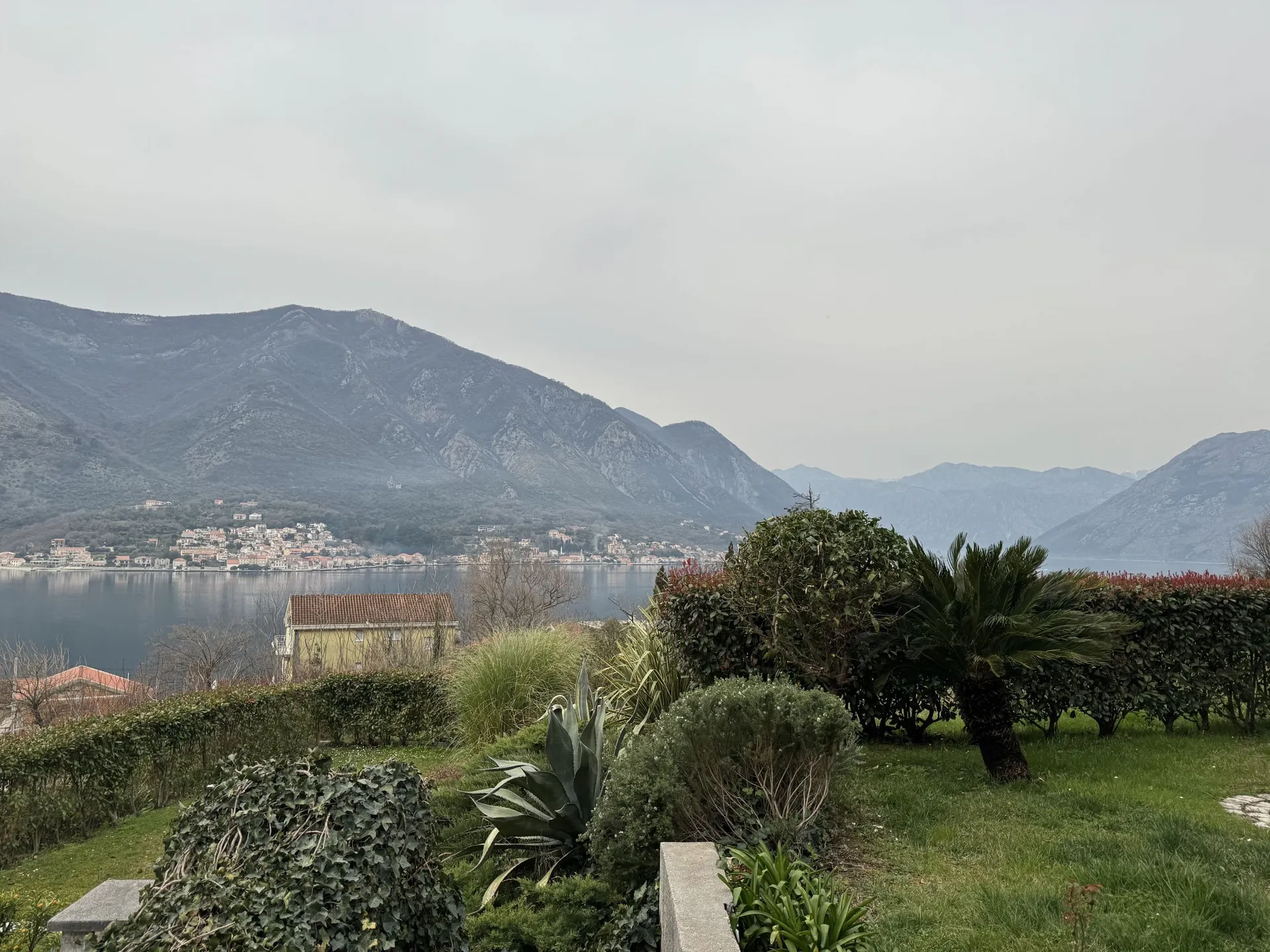 Landscape view of a bay, mountains, and coastal town. Foreground features a manicured garden with palms, bushes, and grass under an overcast sky.