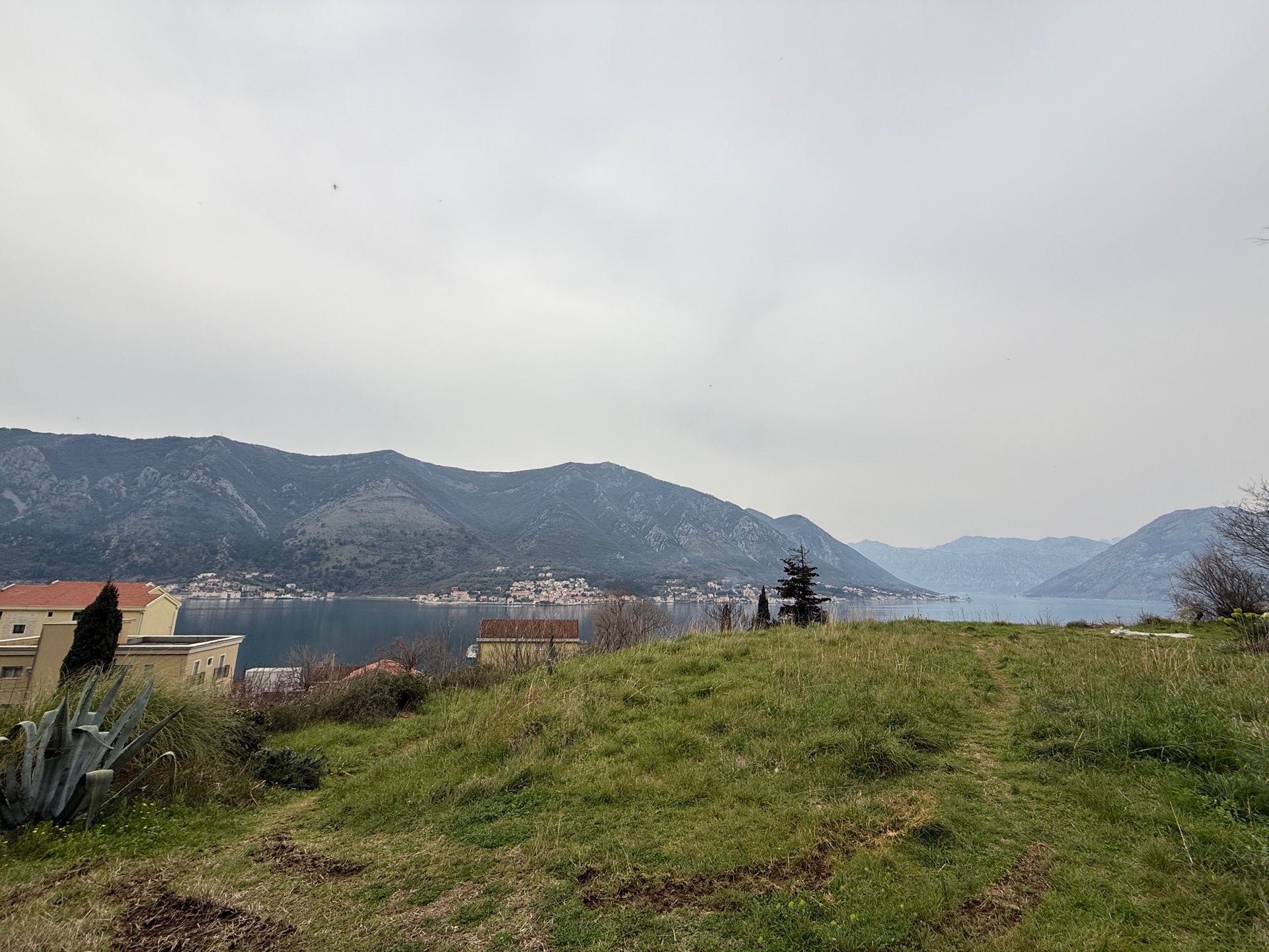 Panoramic view of Kotor Bay and the mountains from Dobrota, Montenegro: a green plot with a house on the coast.