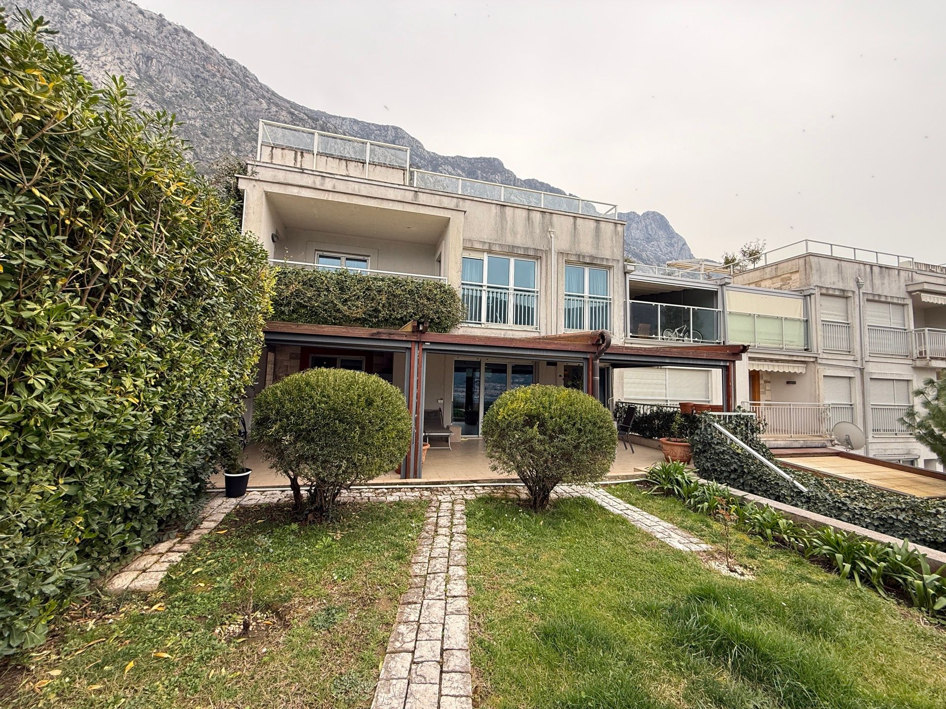 Two-story villa with a terrace and garden in Dobrota, Montenegro: front view of the house and mountain scenery in the background.