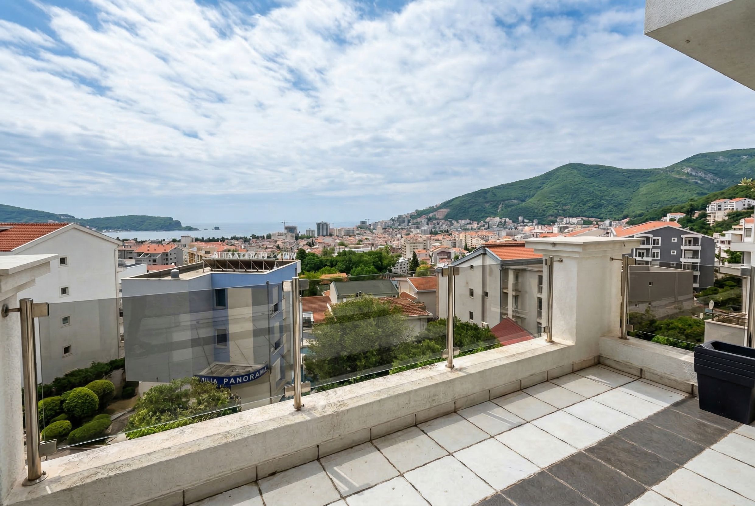 Panoramic view of the sea and mountains from the apartment balcony in Budva, Montenegro, with a modern glass railing.