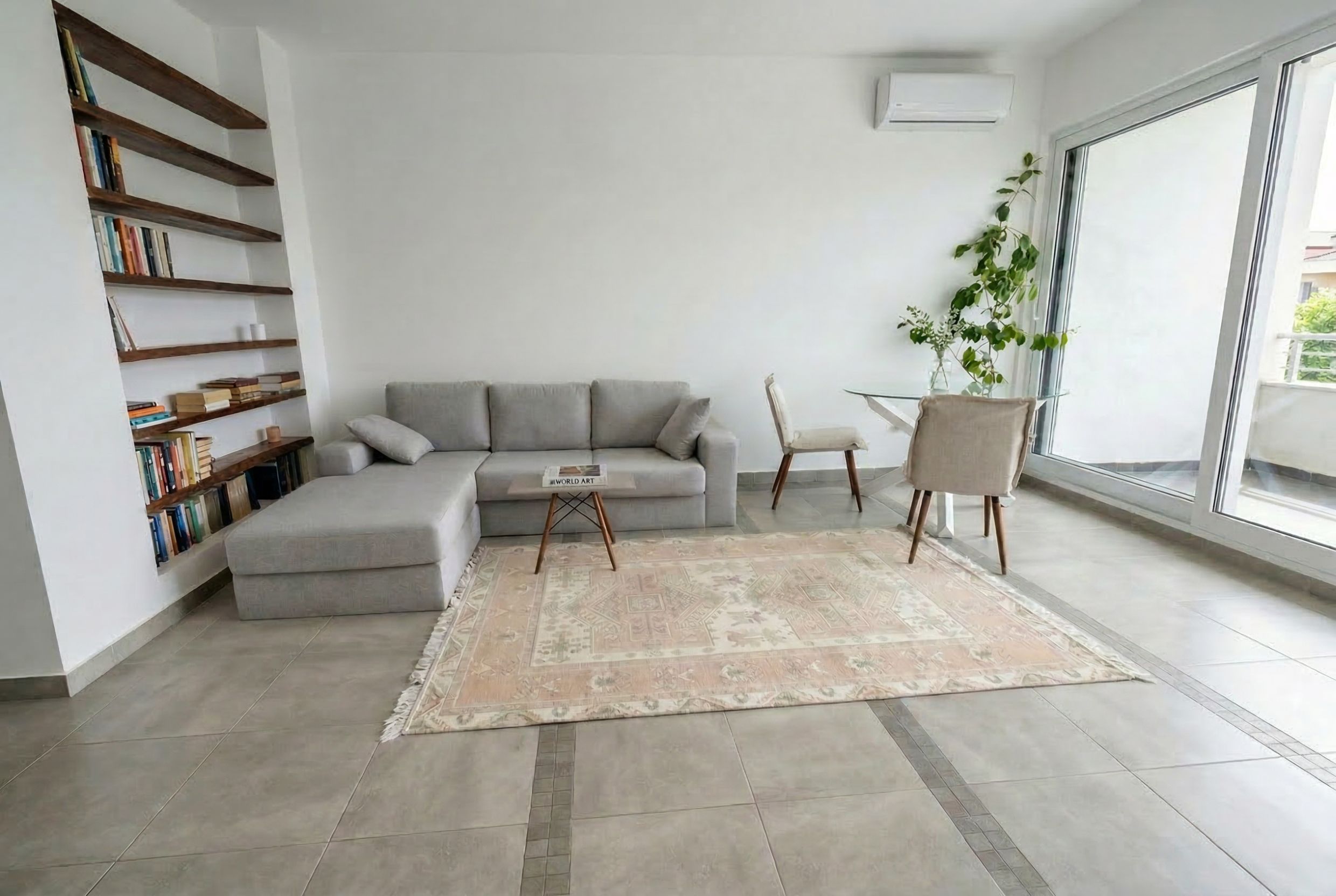 Bright living room in an apartment in Budva, Montenegro: gray corner sofa, dining area, bookshelves, and a panoramic window.