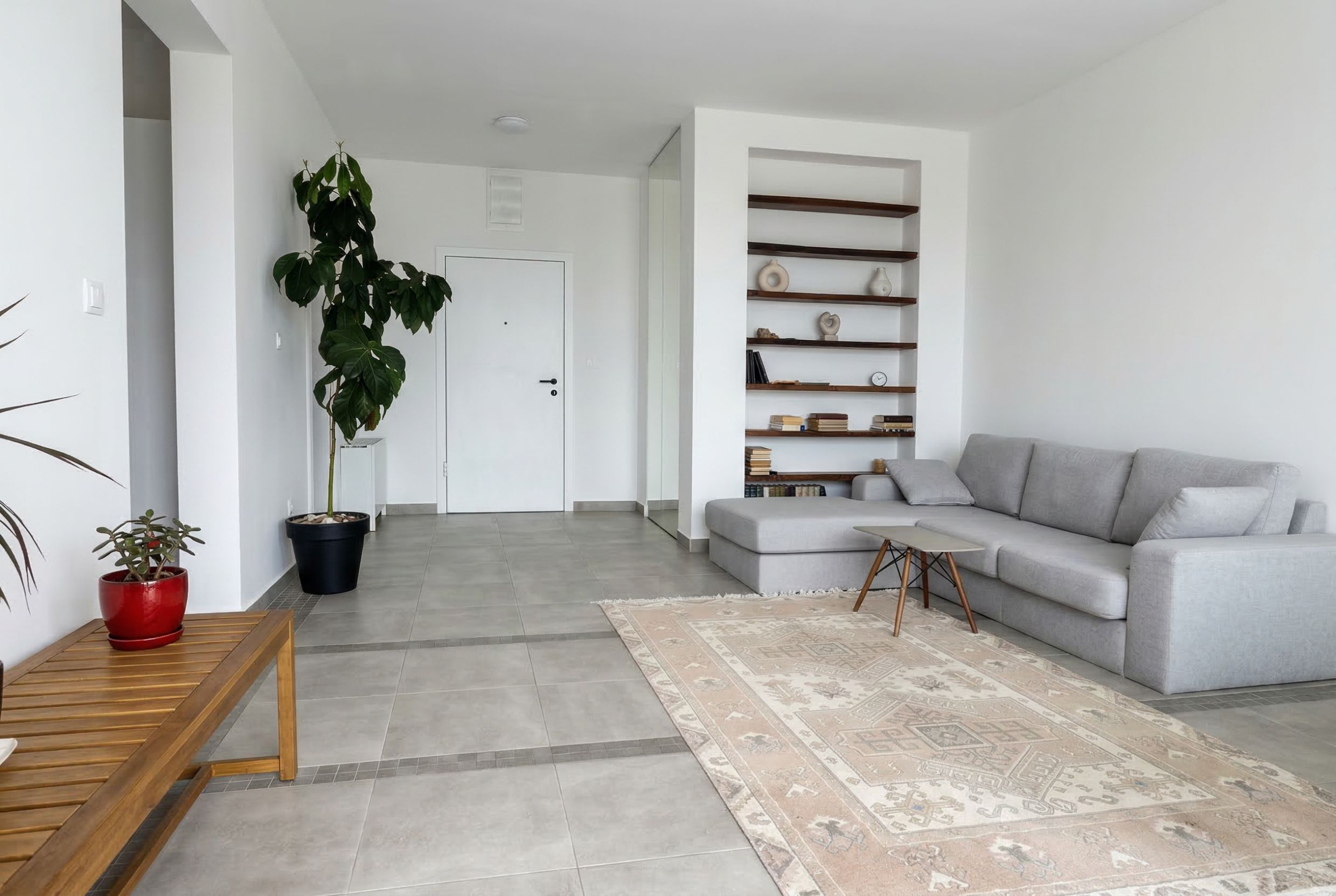 Bright living room in an apartment in Budva, Montenegro: gray sofa, built-in shelves, tiled floor, and houseplants.