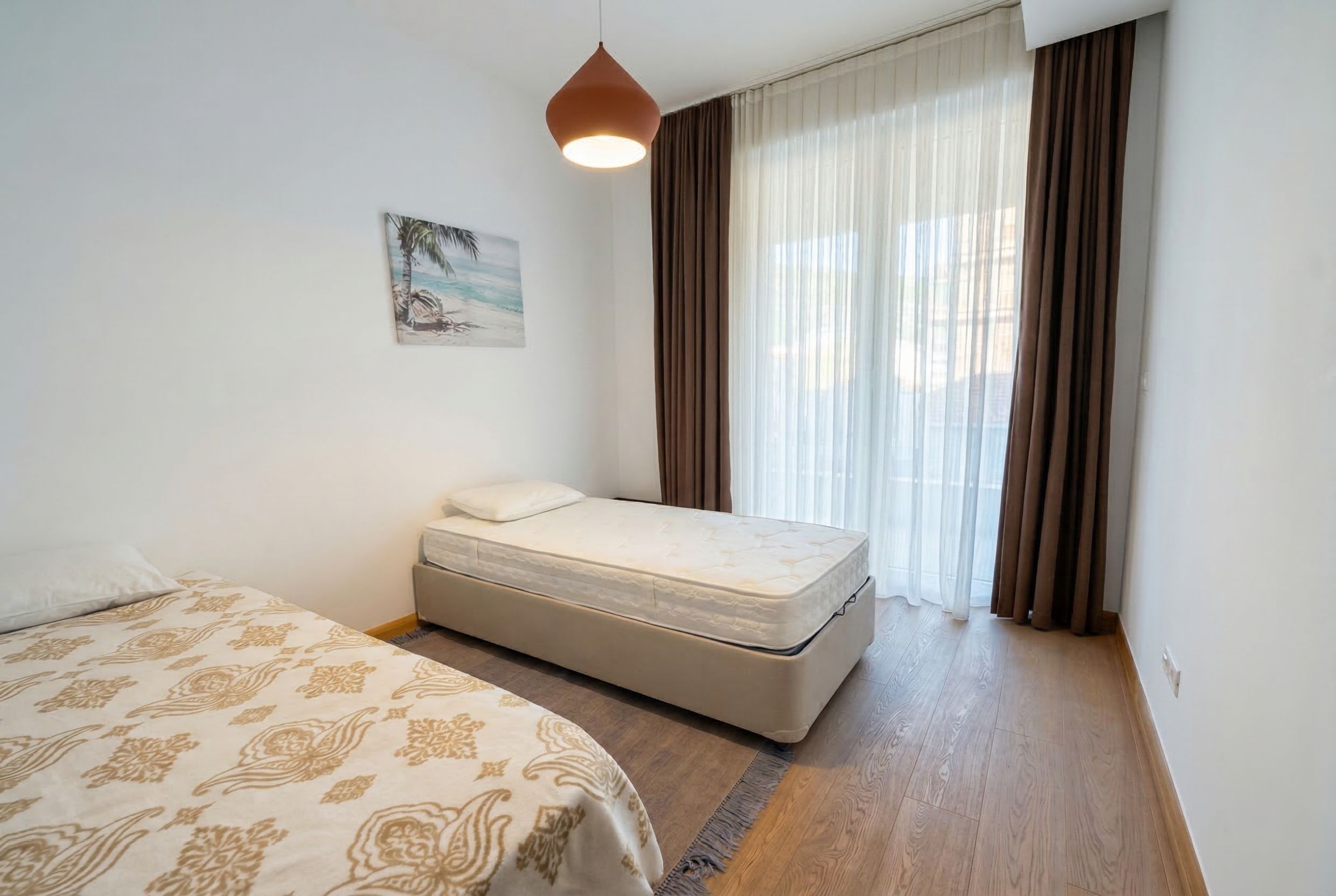 Bright bedroom with two beds, a panoramic window, and brown curtains in an apartment in Budva, Montenegro.