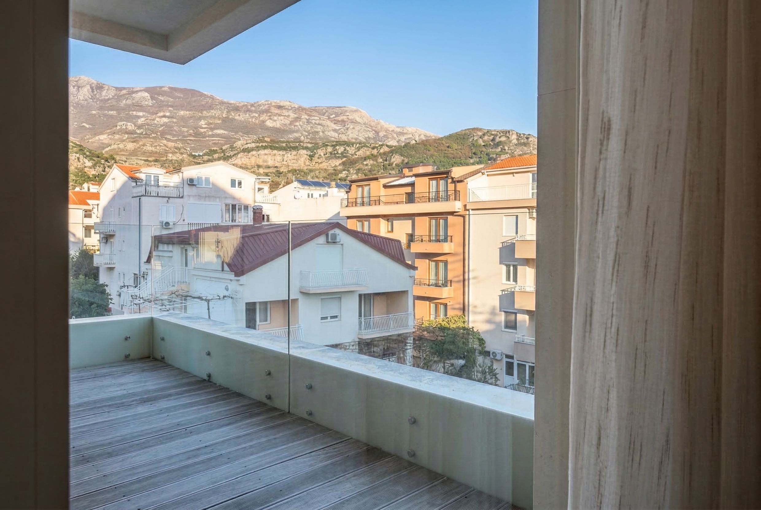 View from a balcony with wooden decking and glass railing of the mountains and residential buildings in Budva, Montenegro.