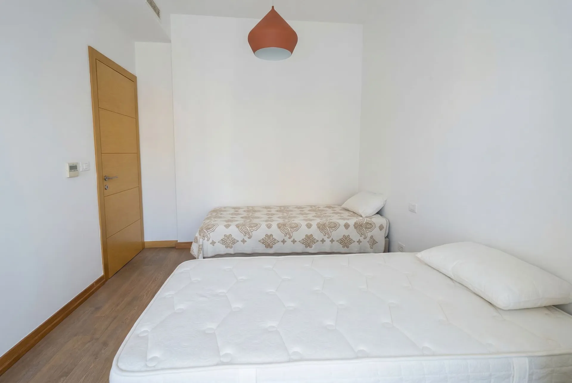 Minimalist bedroom with two beds, a wooden door, and a terracotta pendant lamp against white walls and wood flooring.